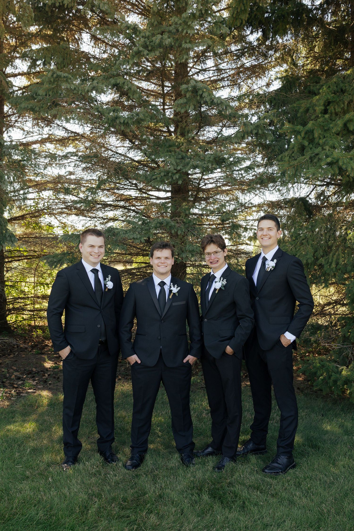 Four groomsmen in black suits with white boutonnieres posing outdoors in front of evergreen trees on a wedding day.
