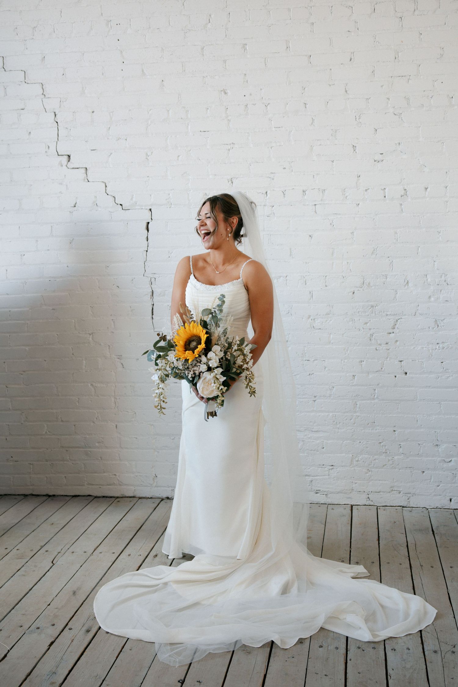 Smiling bride in white spaghetti strap gown with train holds sunflower bouquet against white brick wall.