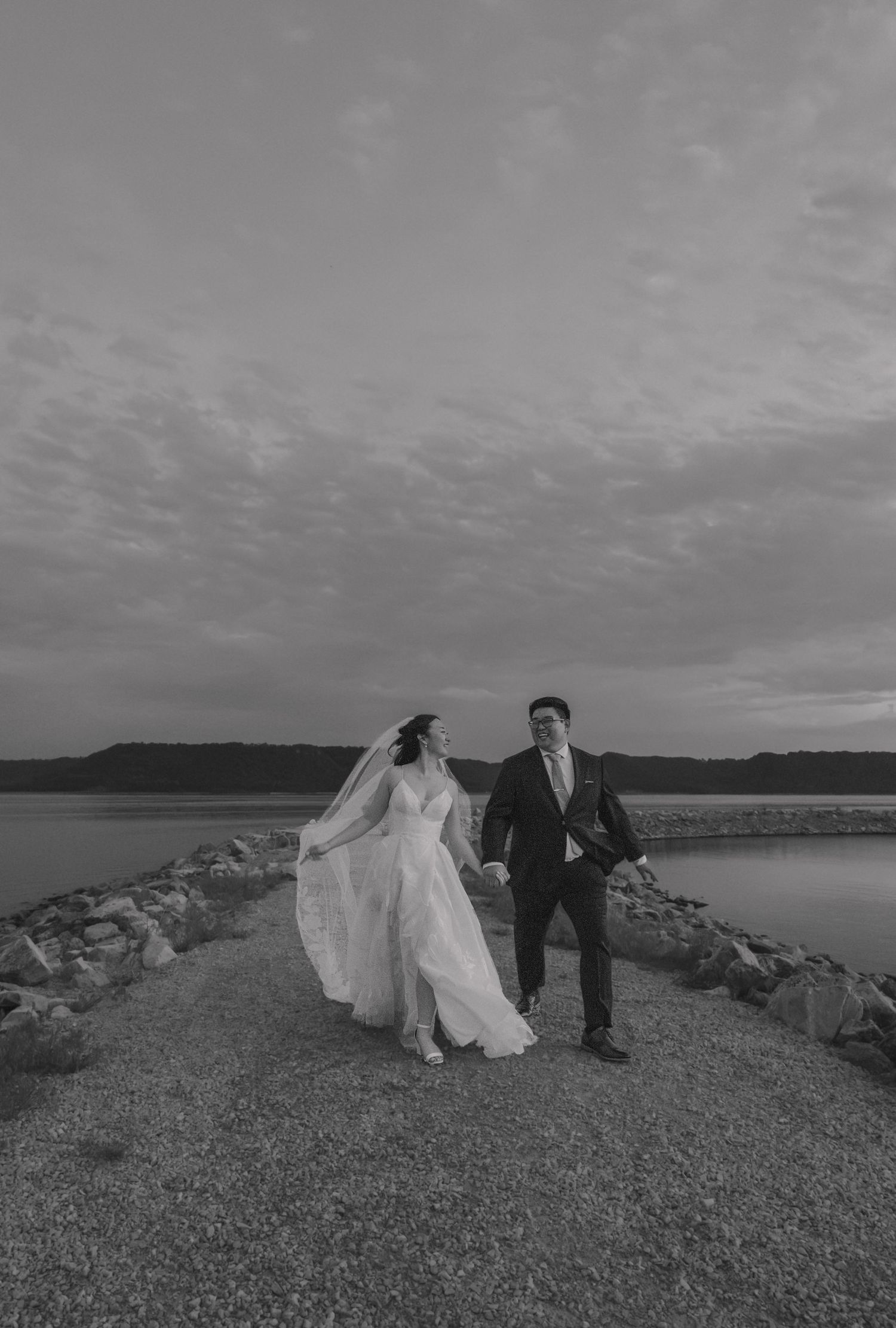 Black and white photo of bride and groom holding hands while walking along a rocky waterfront pier at dusk.