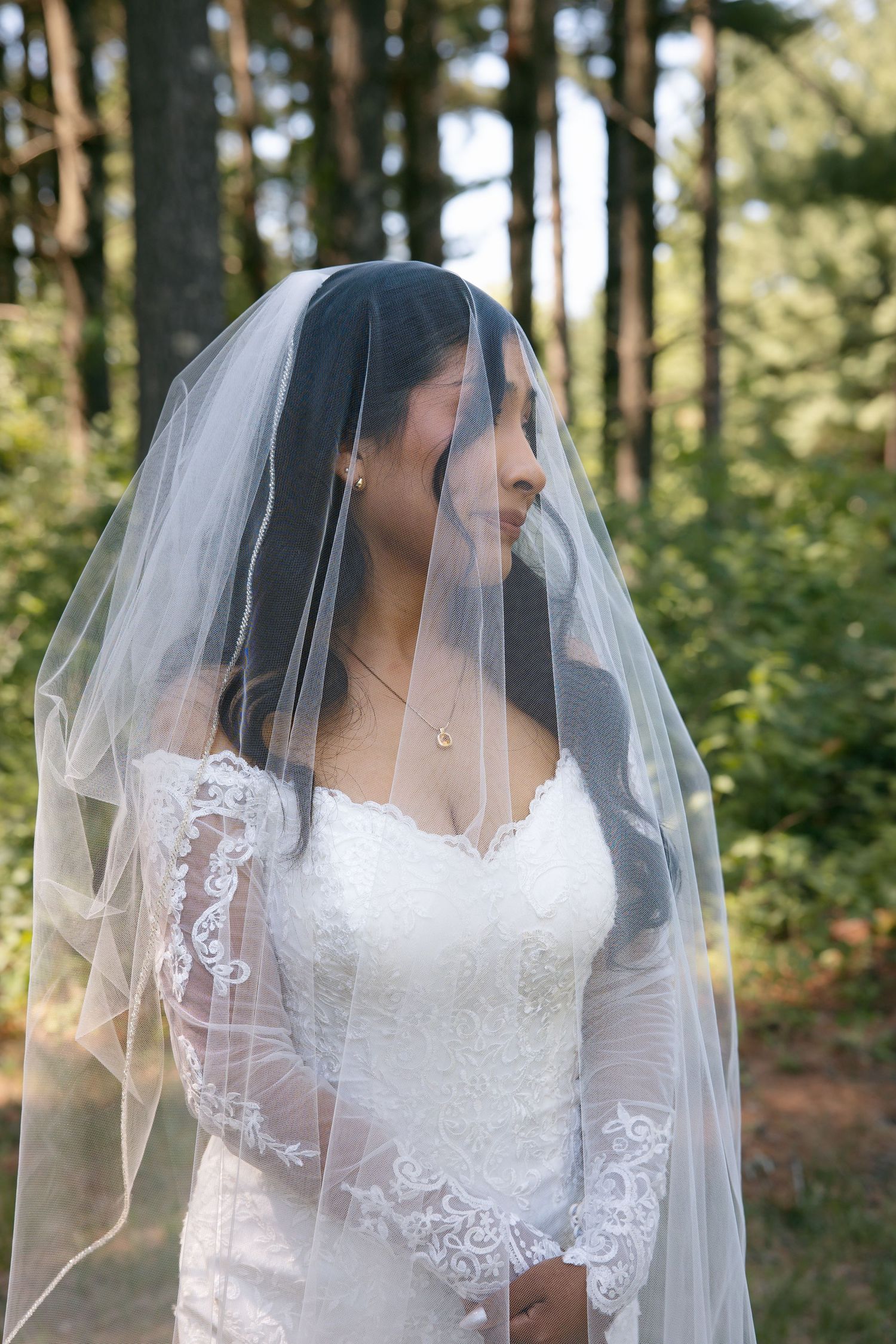 Bride wearing a white lace gown and flowing veil poses outdoors in a forest setting.