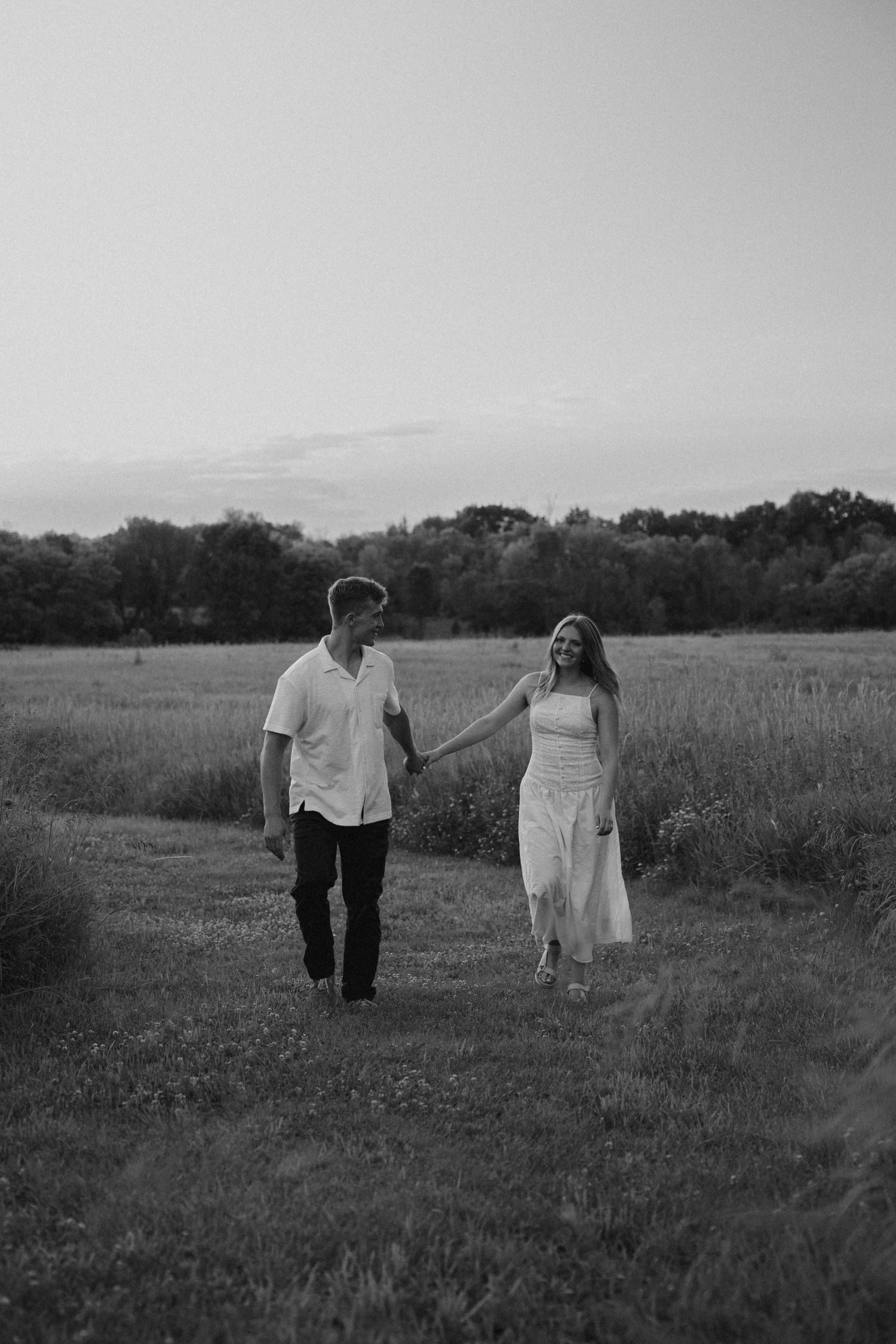 Black and white photo of a couple holding hands and walking through a lush green field at sunset.