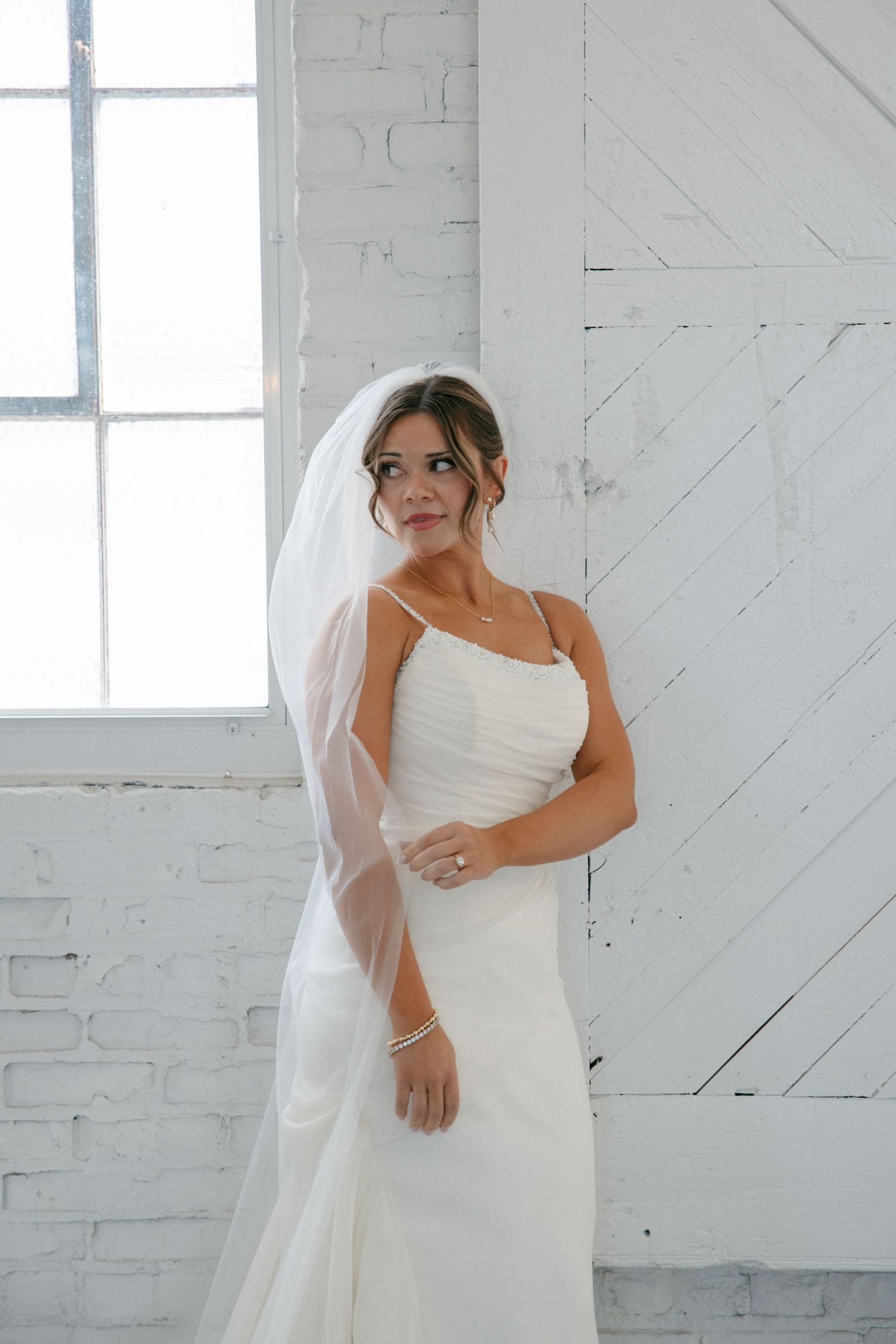 Bride in white spaghetti strap gown and veil poses gracefully in bright white brick room.