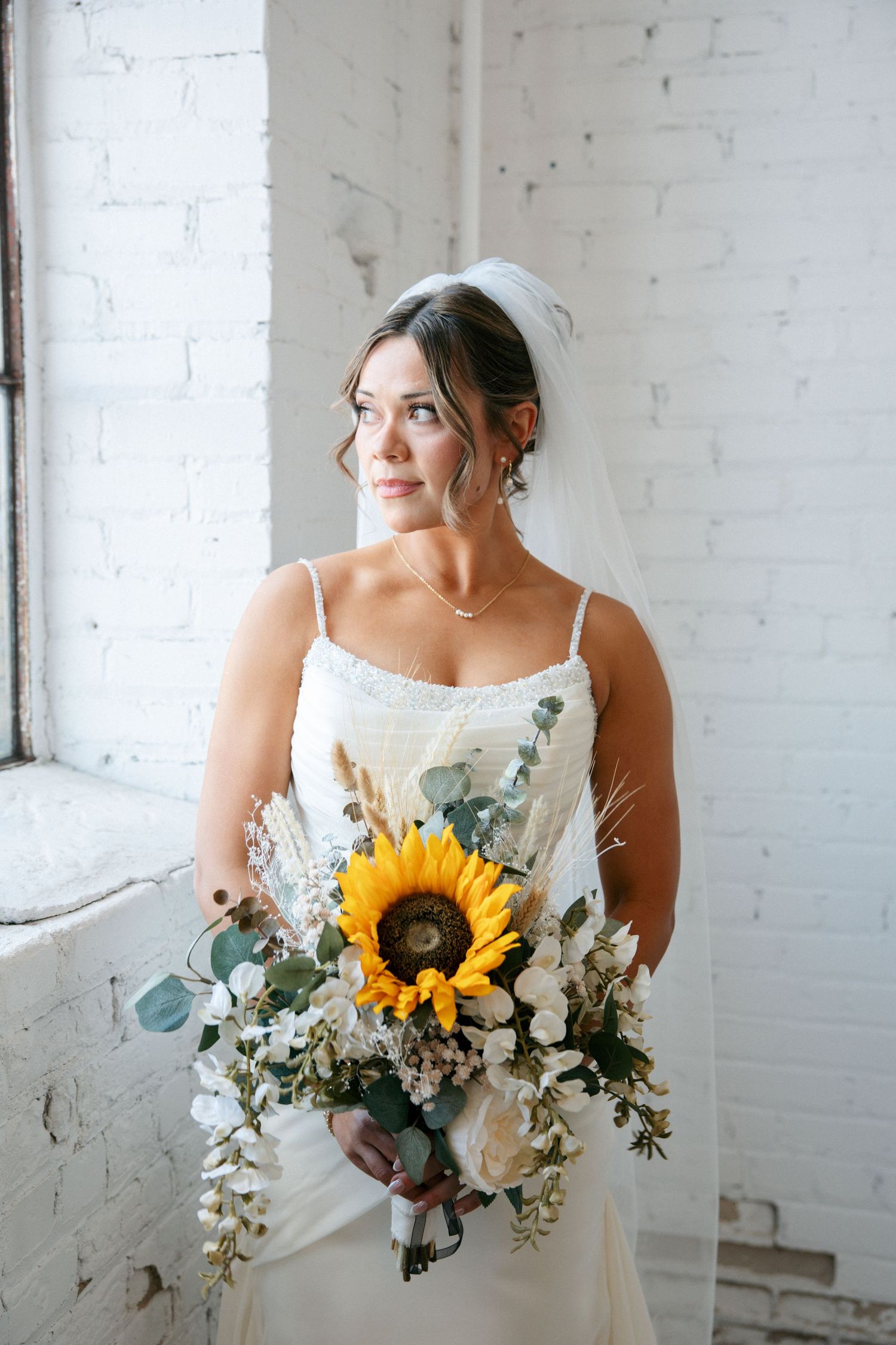 Bride in white dress holding sunflower bouquet with eucalyptus, posing by a white brick wall window.