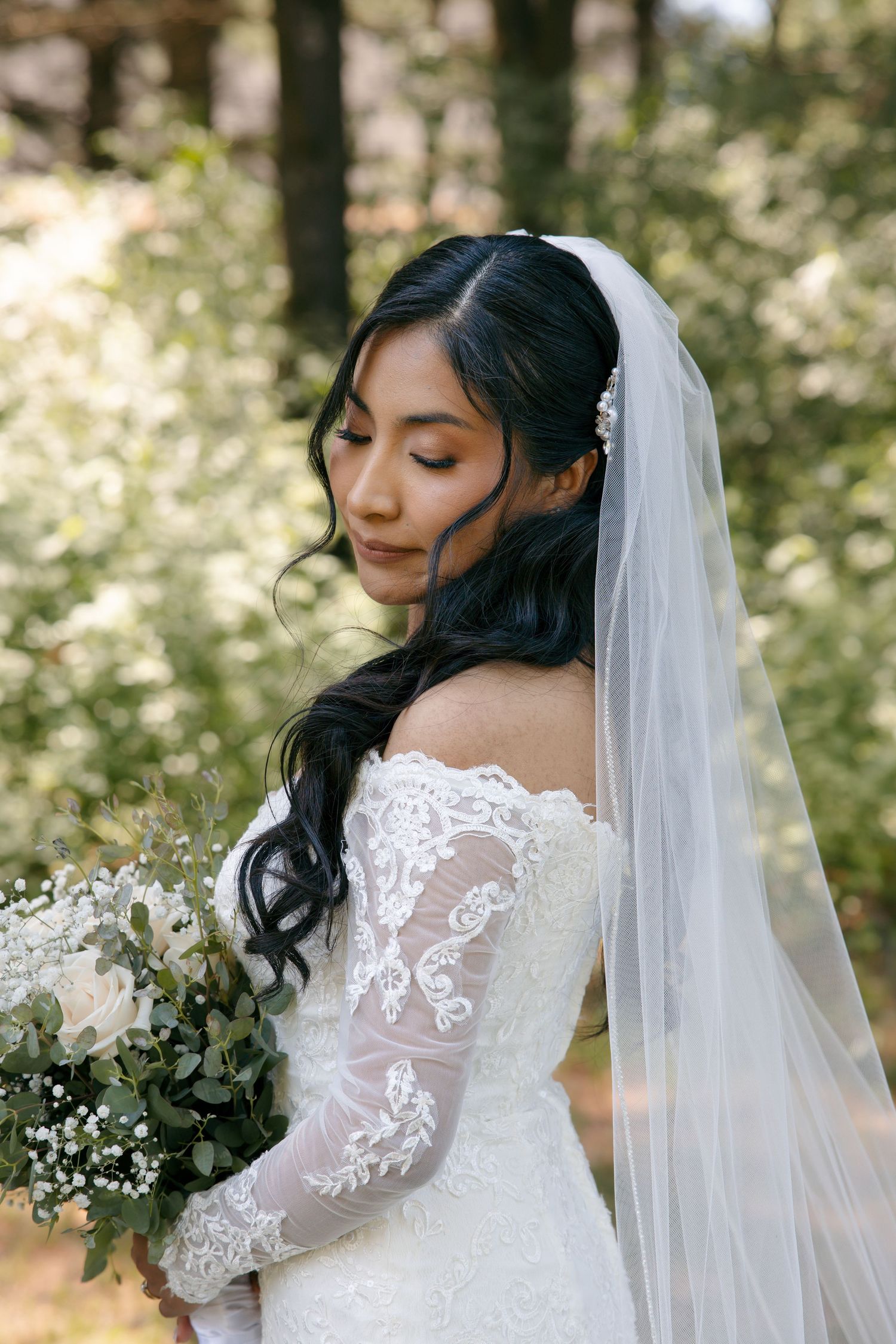 Bride in lace off-shoulder gown and veil holds white bouquet, posing outdoors among green trees.
