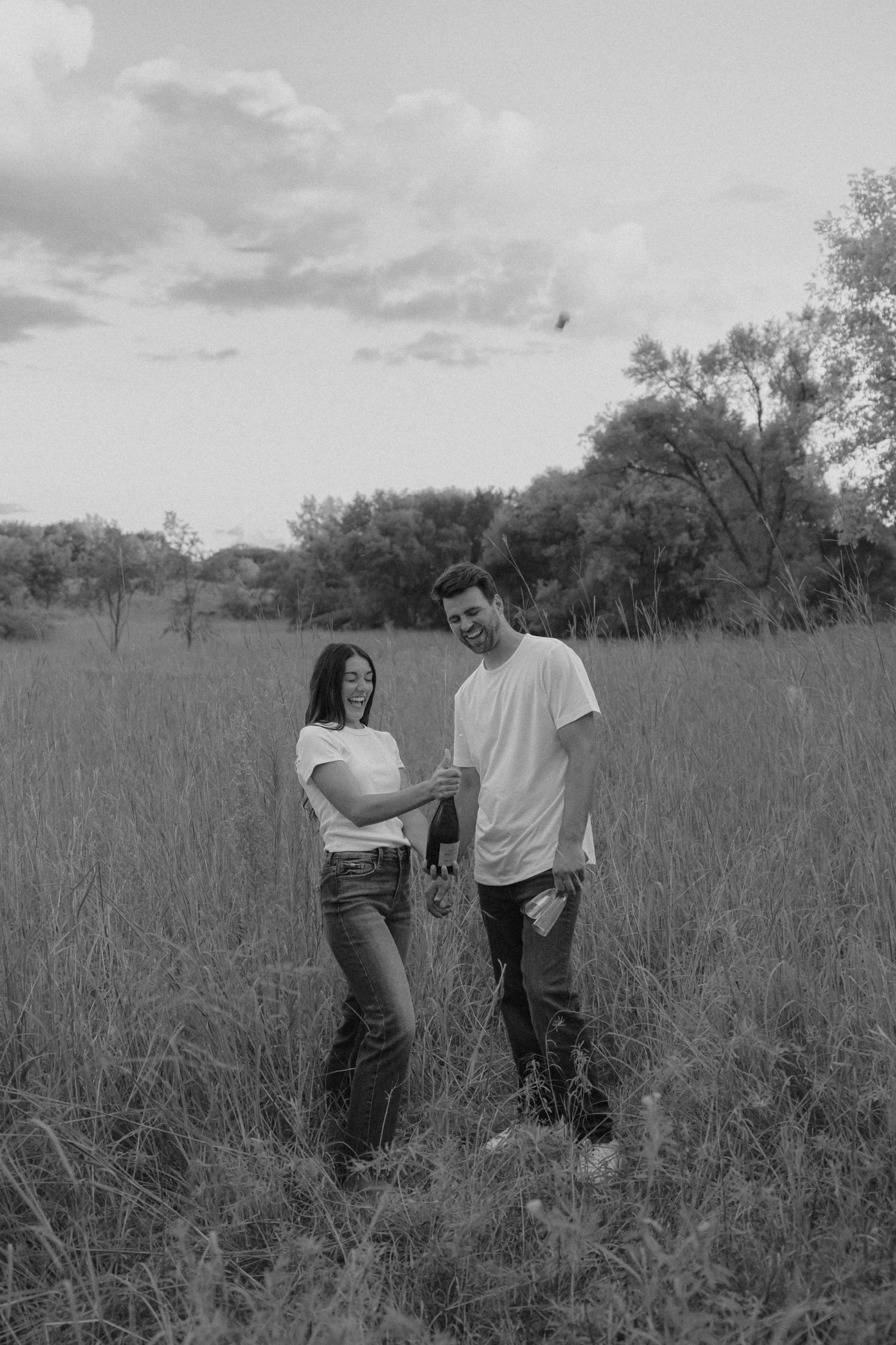 Black and white photo of a couple holding hands and smiling in a grassy field with trees in the background.