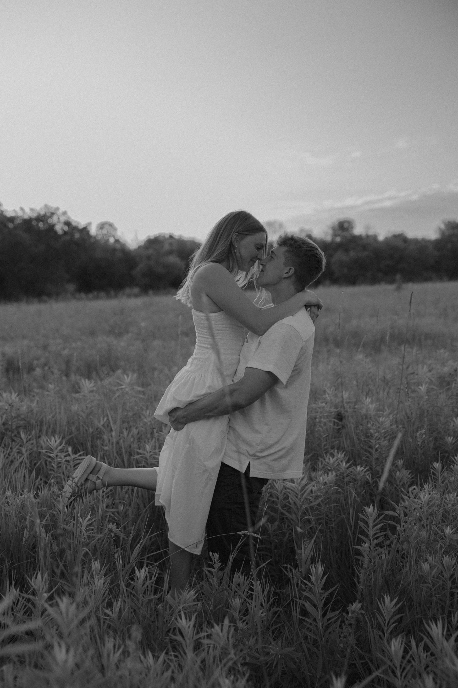 Black and white photo of a couple kissing in a field at sunset, man lifting woman in a romantic outdoor setting.