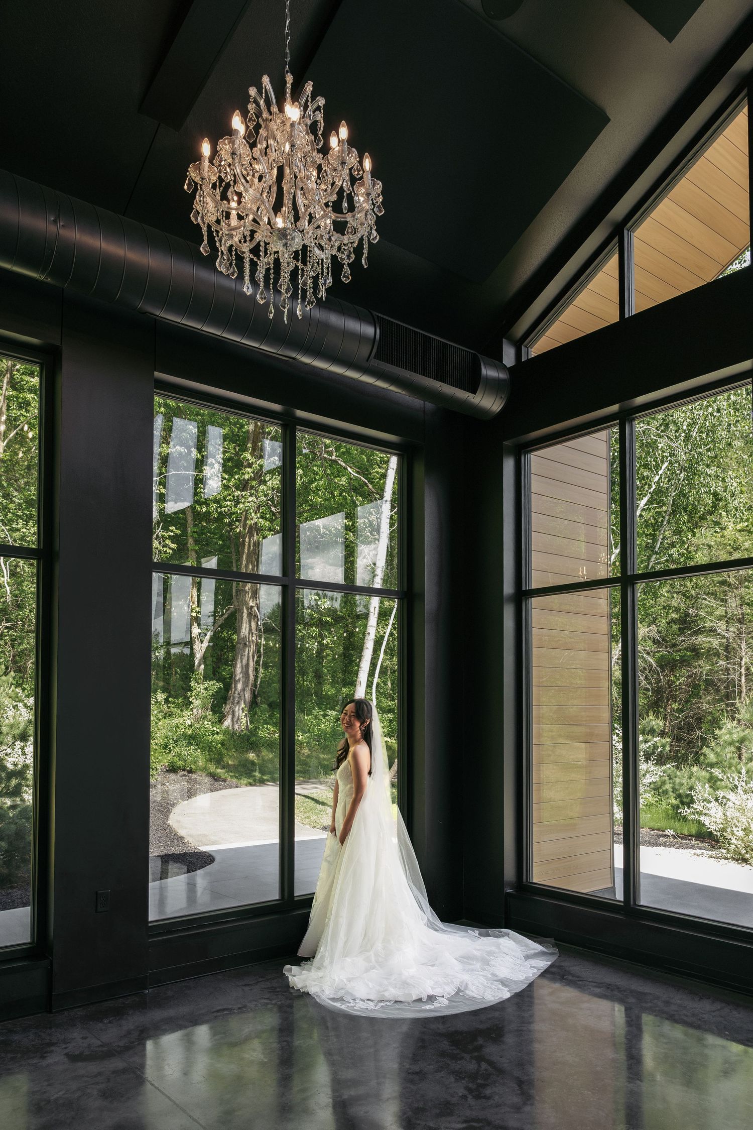 Bride in a flowing white gown and long veil stands beneath a crystal chandelier in a modern venue.