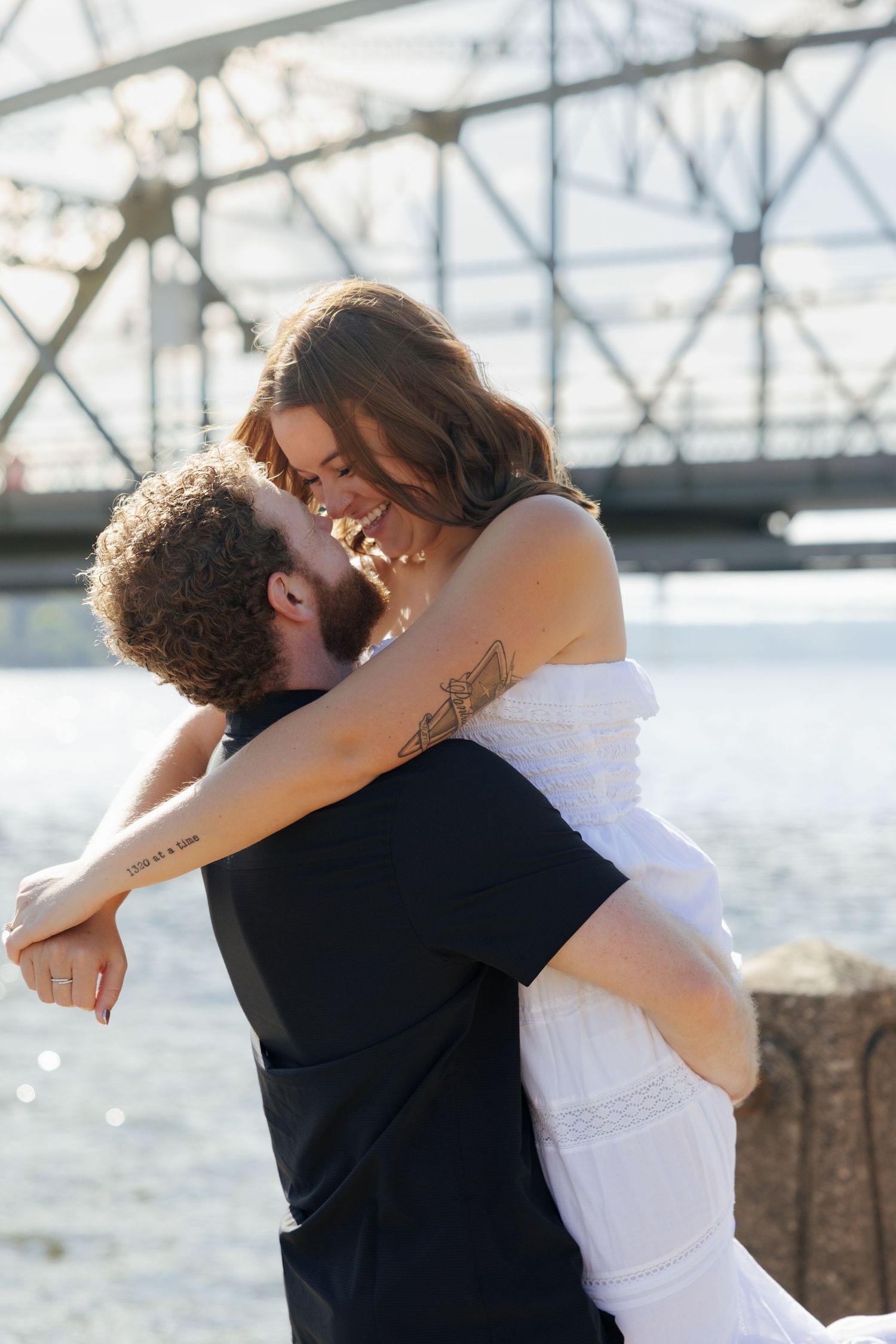A groom lifts his smiling bride in a white dress near a waterfront bridge on their wedding day.