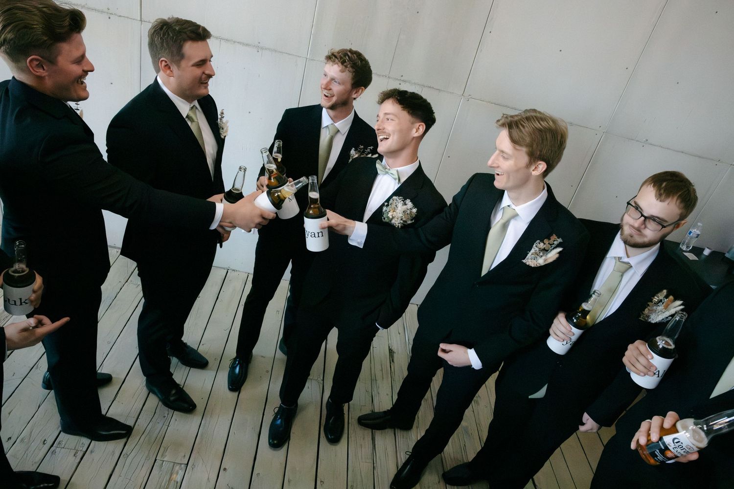 Groomsmen in black suits laughing and toasting with champagne bottles before the wedding ceremony.