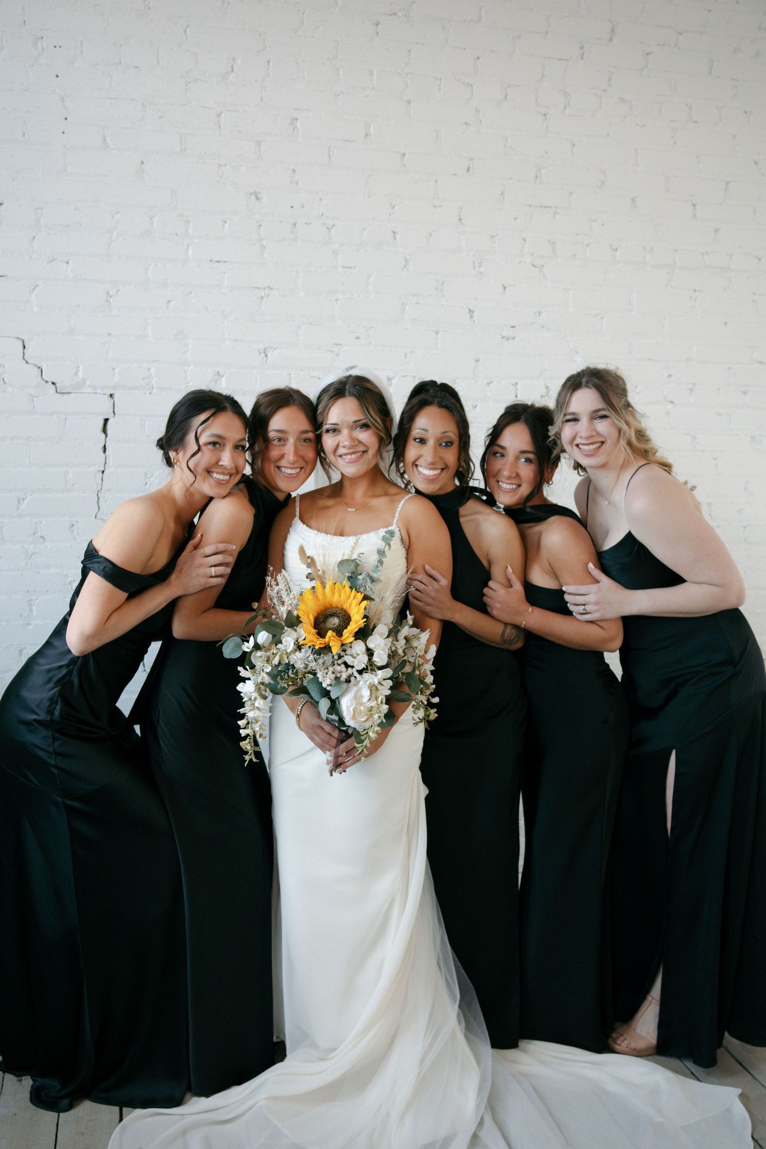 Smiling bride holding sunflower bouquet surrounded by five bridesmaids in black gowns against white brick.