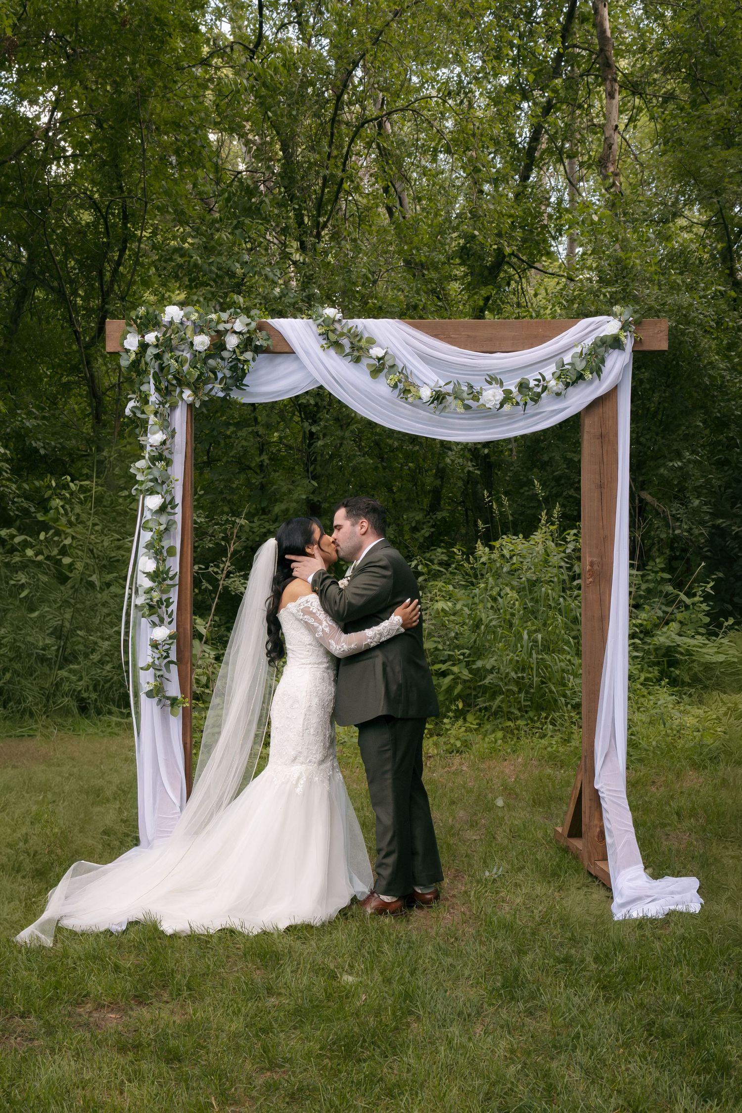 Bride and groom share first kiss under decorated wooden arch with white draping and greenery in outdoor garden wedding ceremony.