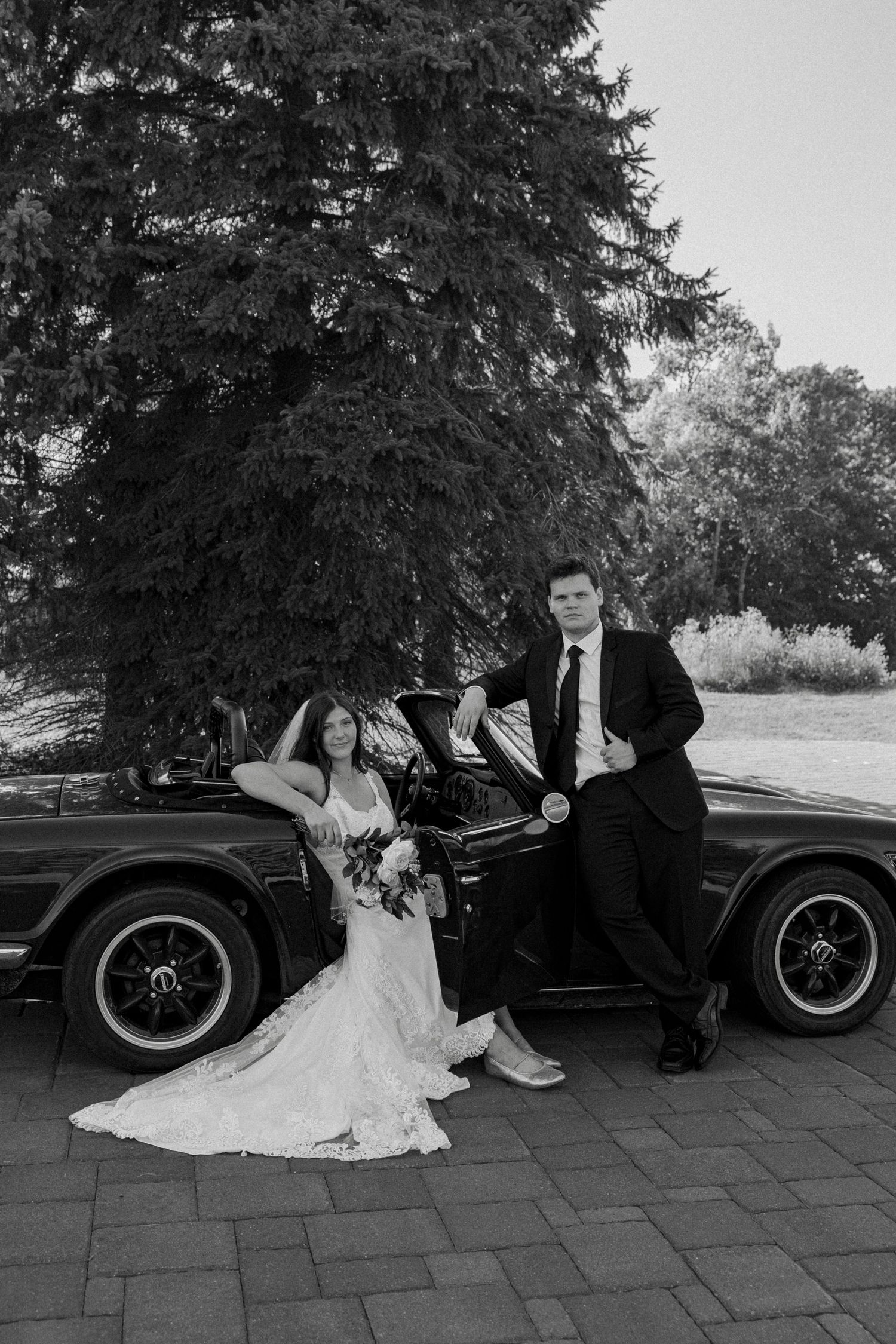 Black and white photo of bride and groom posing elegantly beside a vintage convertible car outdoors.
