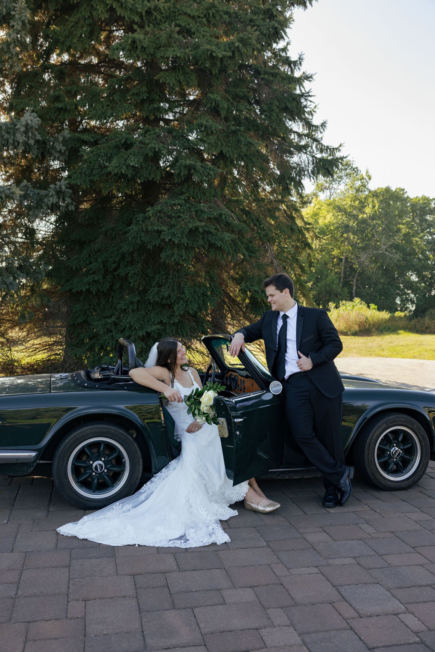 Bride and groom posing beside vintage green convertible car on sunny wedding day.