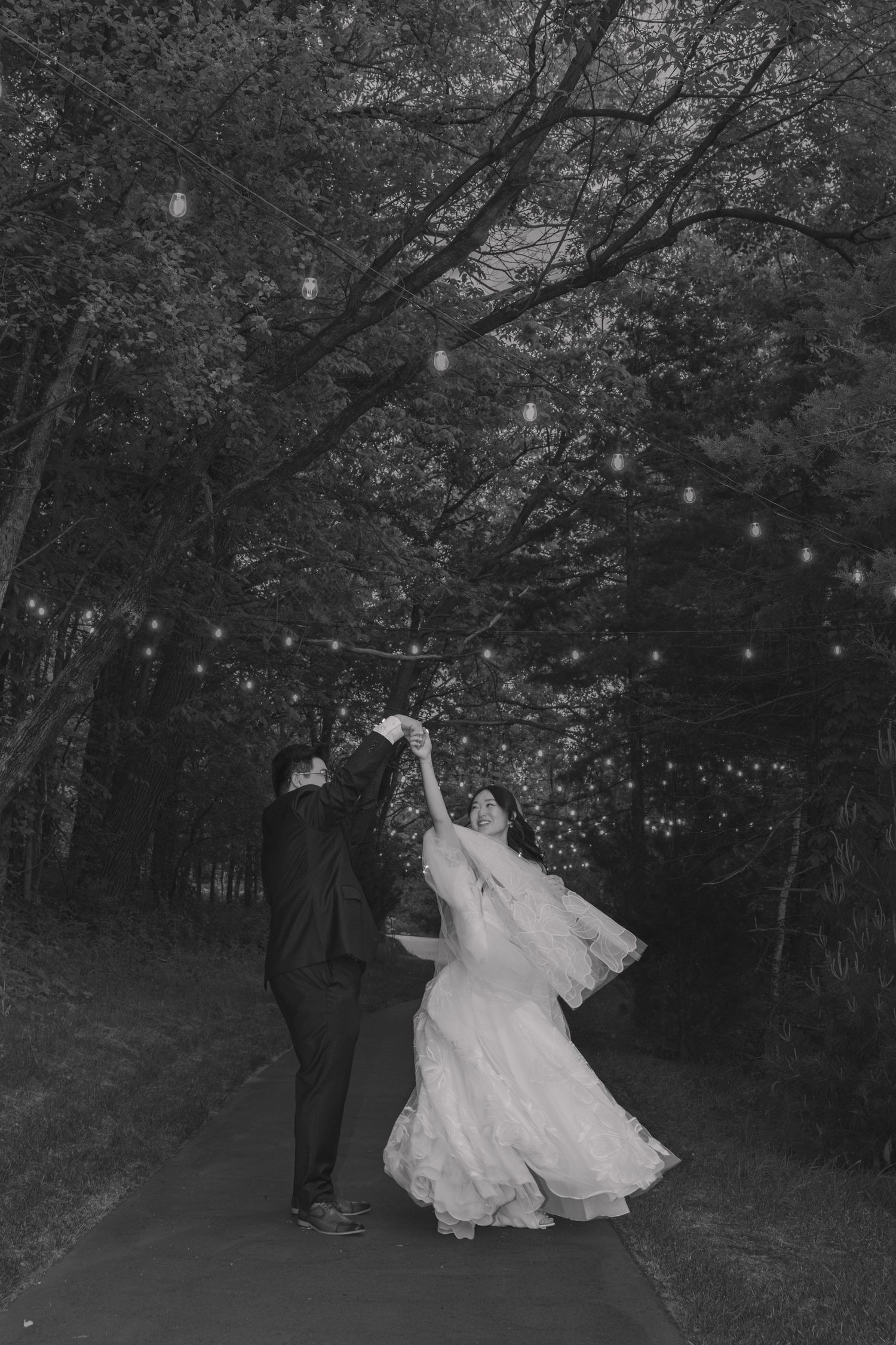 Black and white photo of a bride and groom dancing on a tree-lined path with string lights overhead.