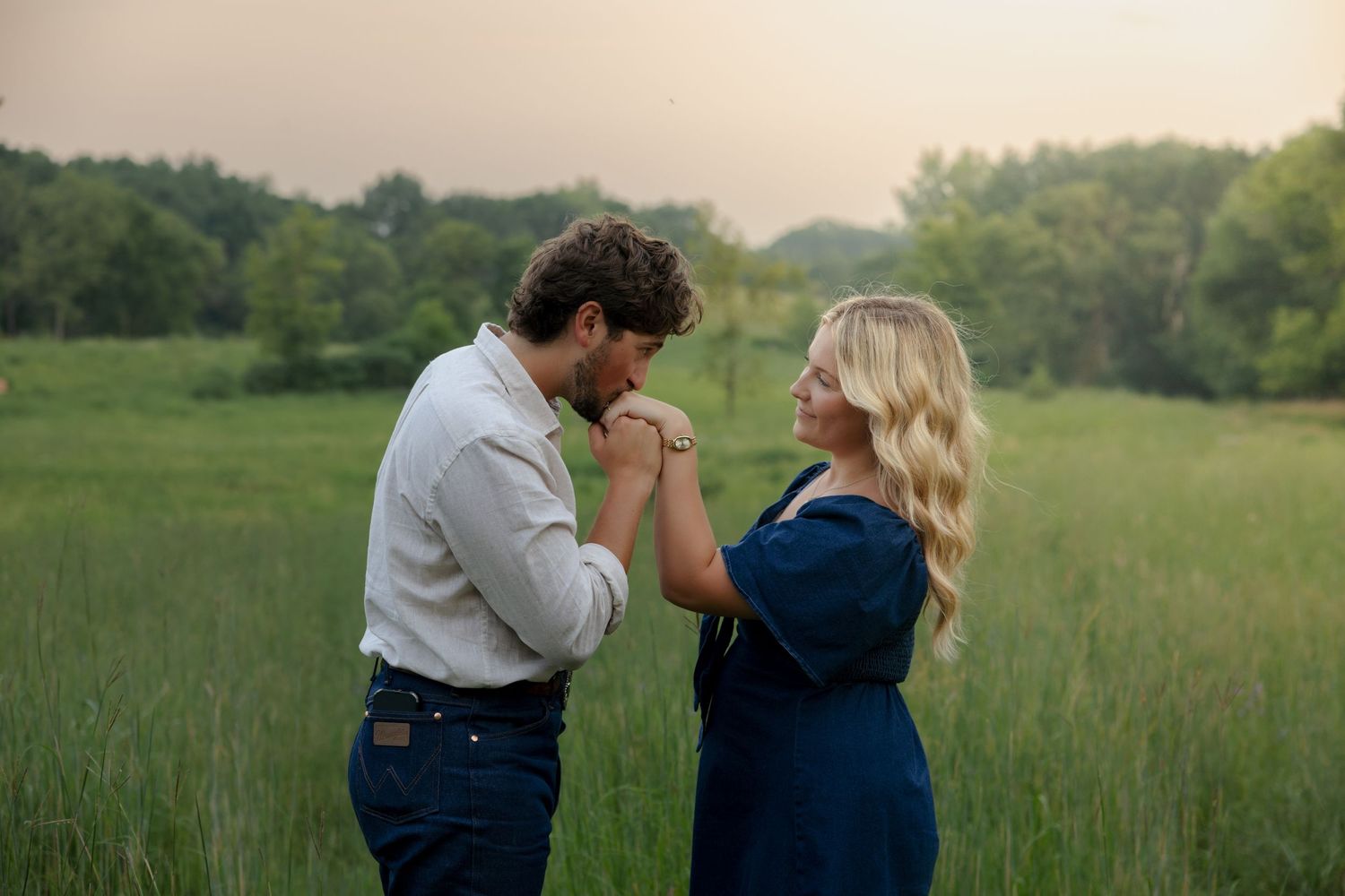 Couple sharing a romantic moment in a green meadow at sunset, man kissing woman's hand.