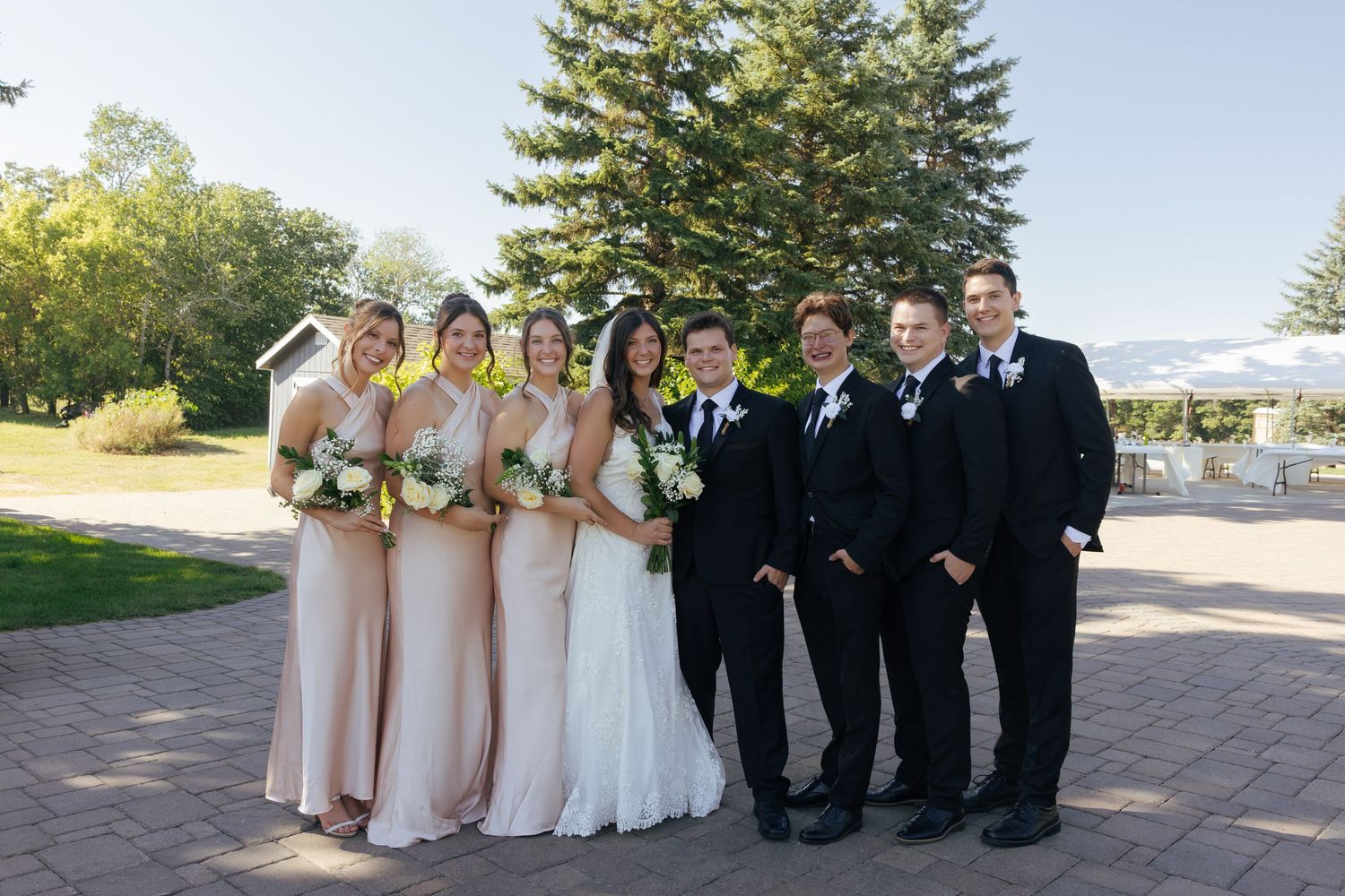 Wedding party posing outdoors, bridesmaids in champagne gowns and groomsmen in black suits flanking bride and groom.