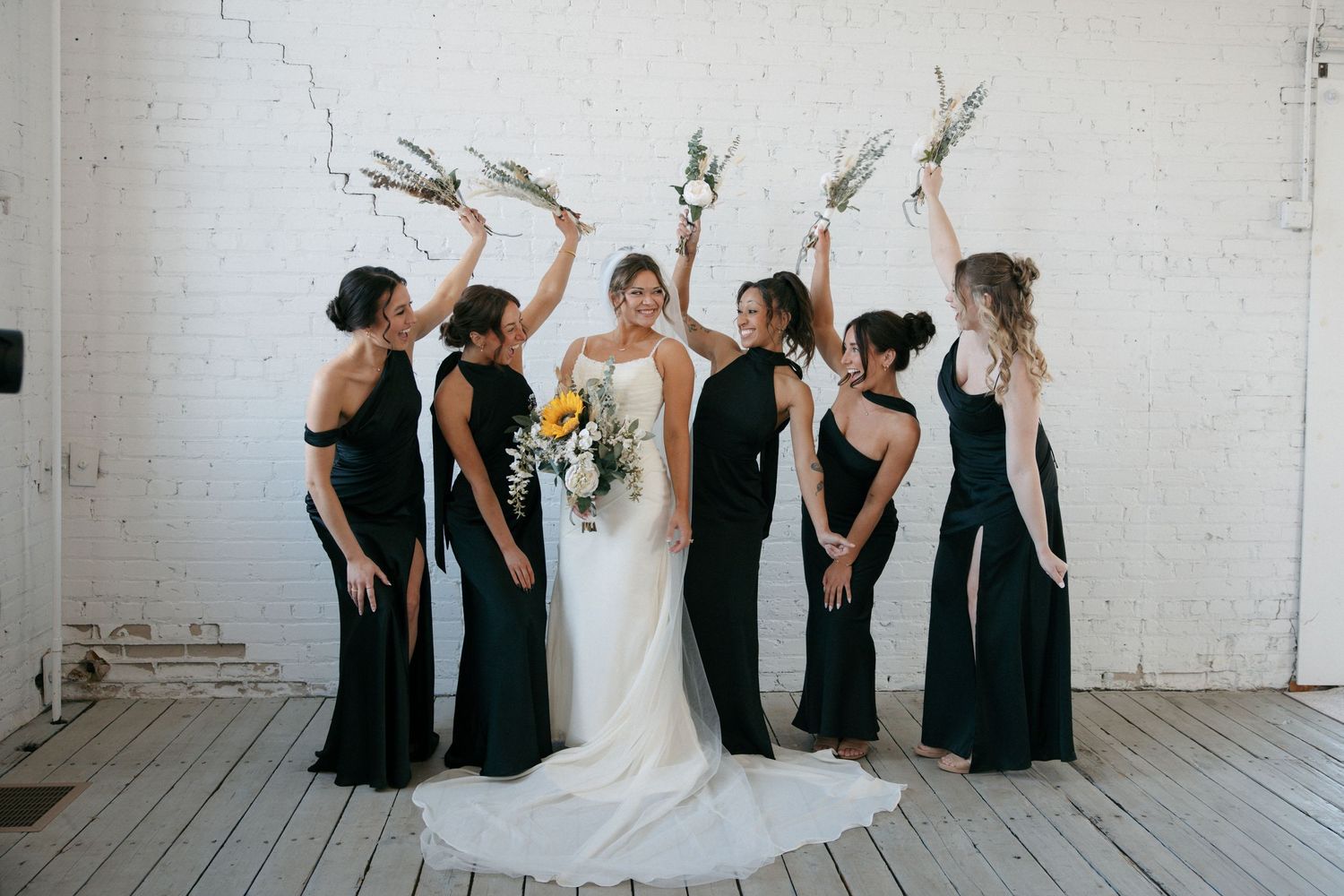 Bride in white gown with six bridesmaids in black dresses raising bouquets in an industrial white brick venue.