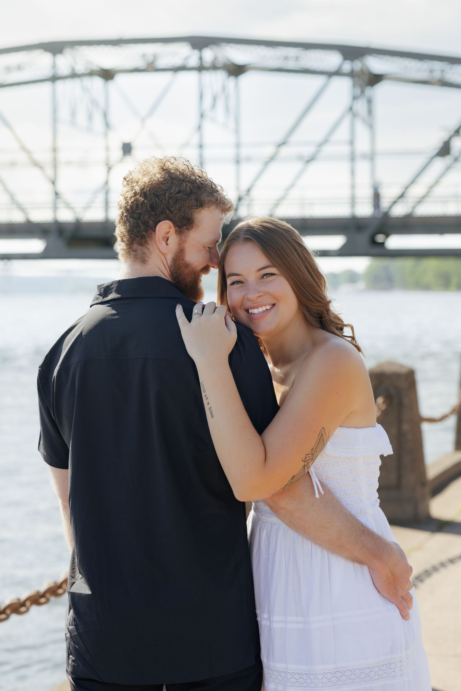 Happy couple embracing near a river with a steel bridge backdrop, woman in white dress smiling warmly.
