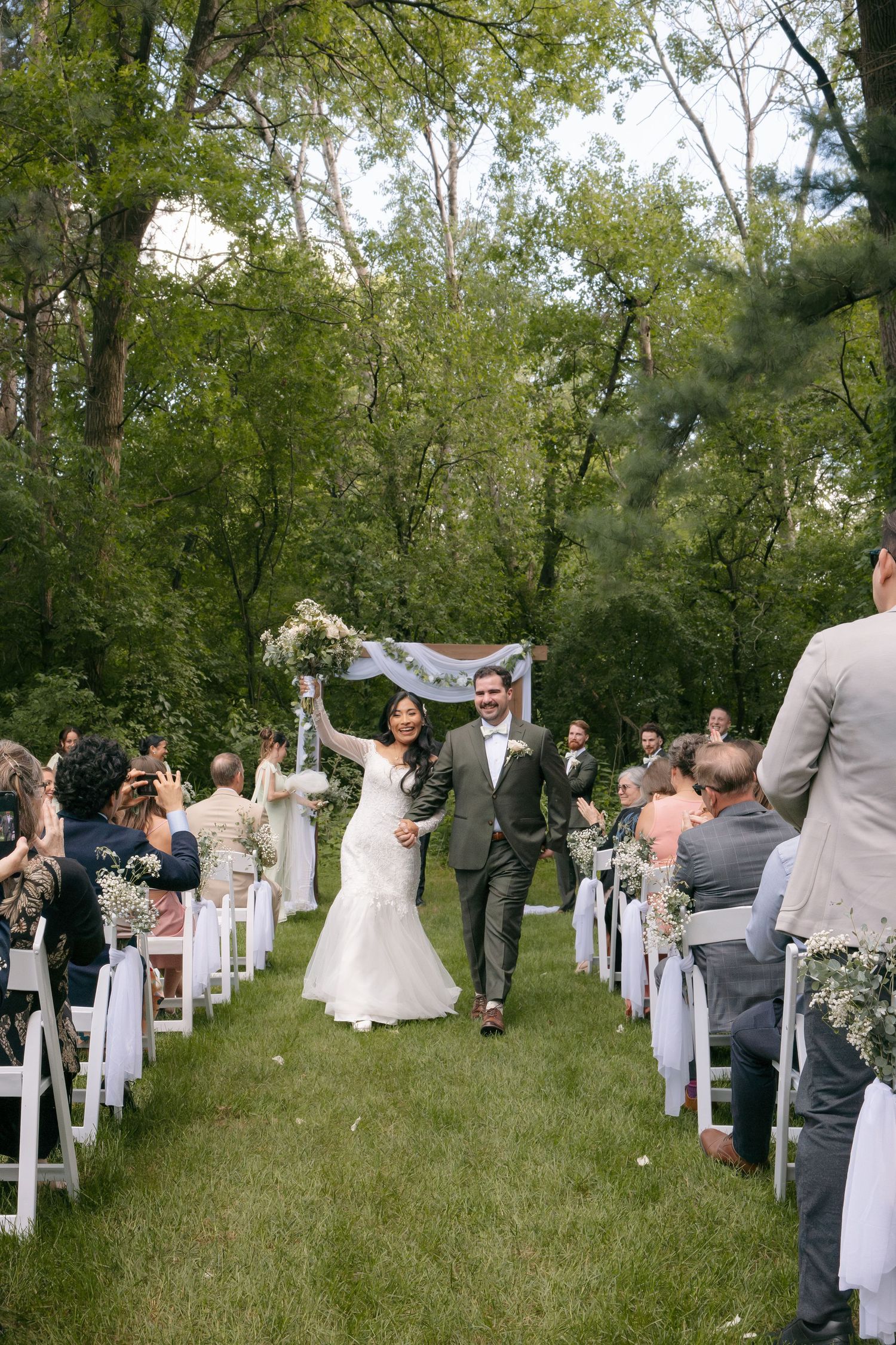 Bride and groom exit wedding ceremony down grass aisle lined with white chairs and guests.