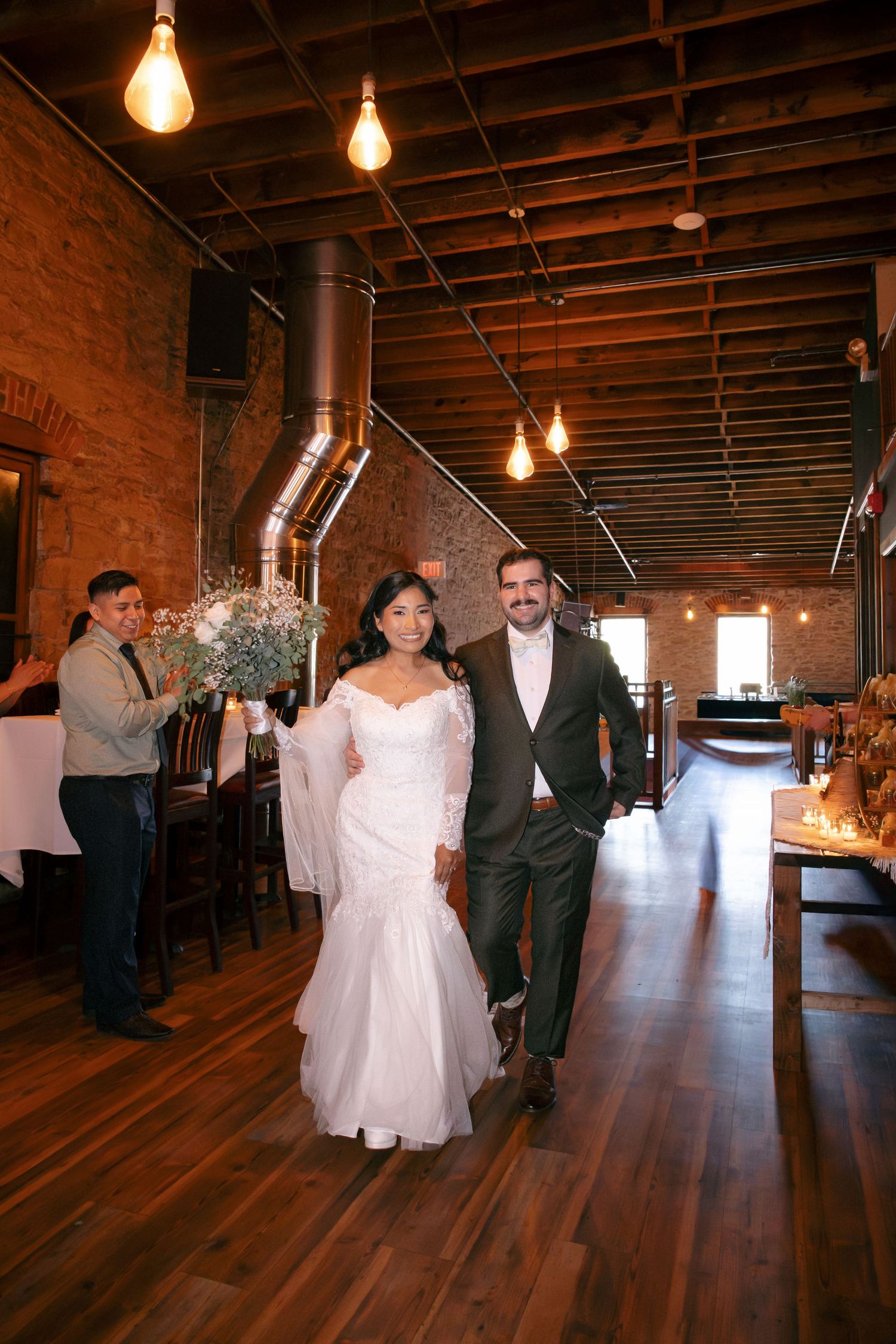 Bride and groom walk hand-in-hand through a rustic brick-walled wedding venue with warm Edison bulb lighting.
