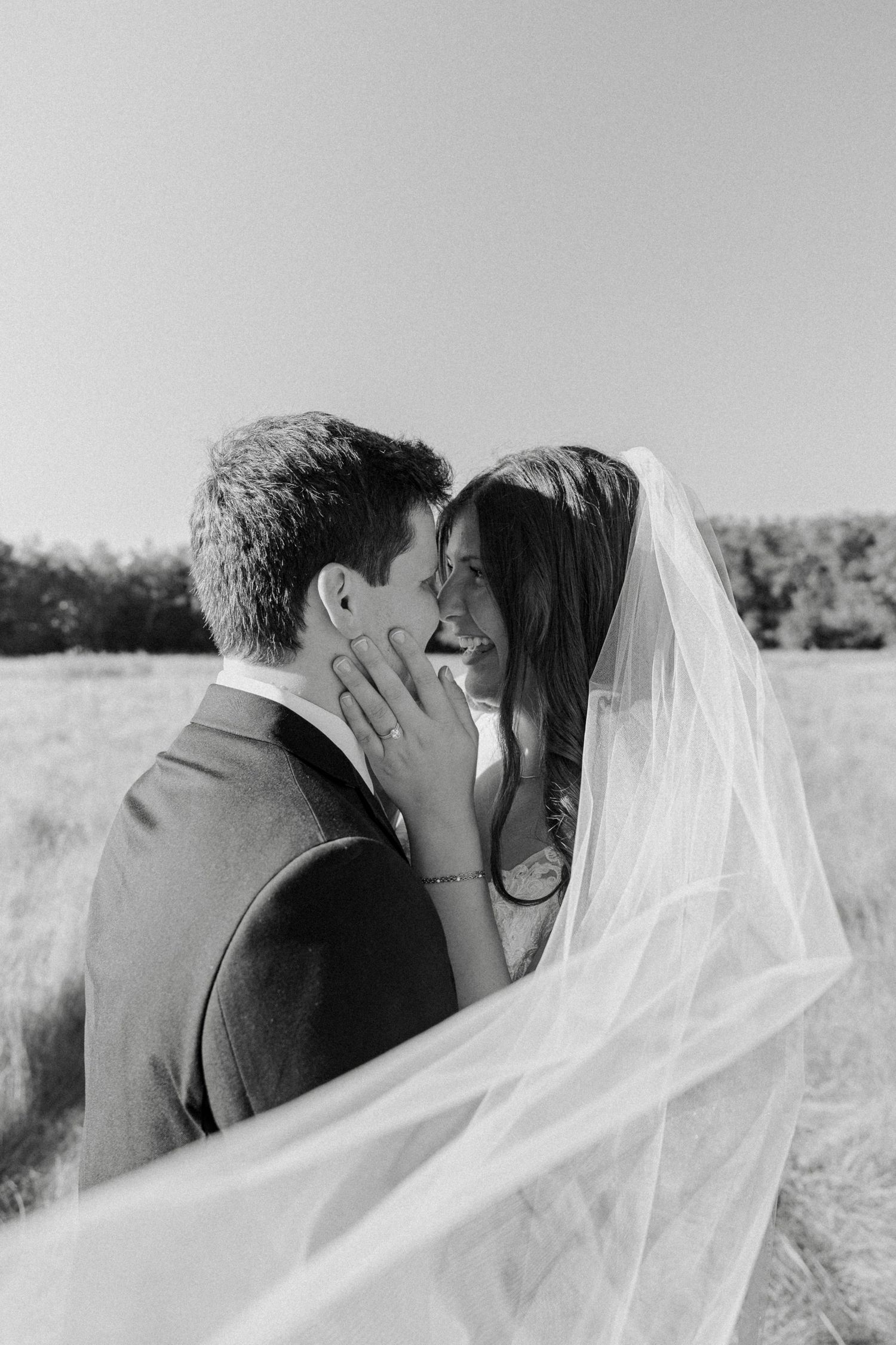 Black and white romantic photo of bride and groom nearly kissing under a flowing veil in an open field.