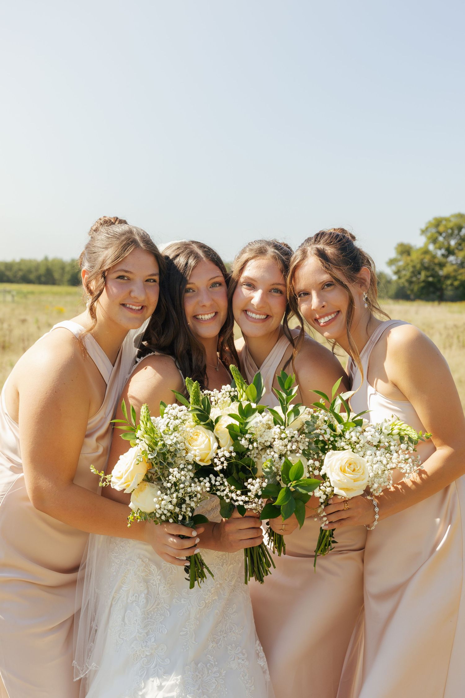 Bride and three bridesmaids in champagne dresses holding white rose bouquets smiling outdoors in sunny field.