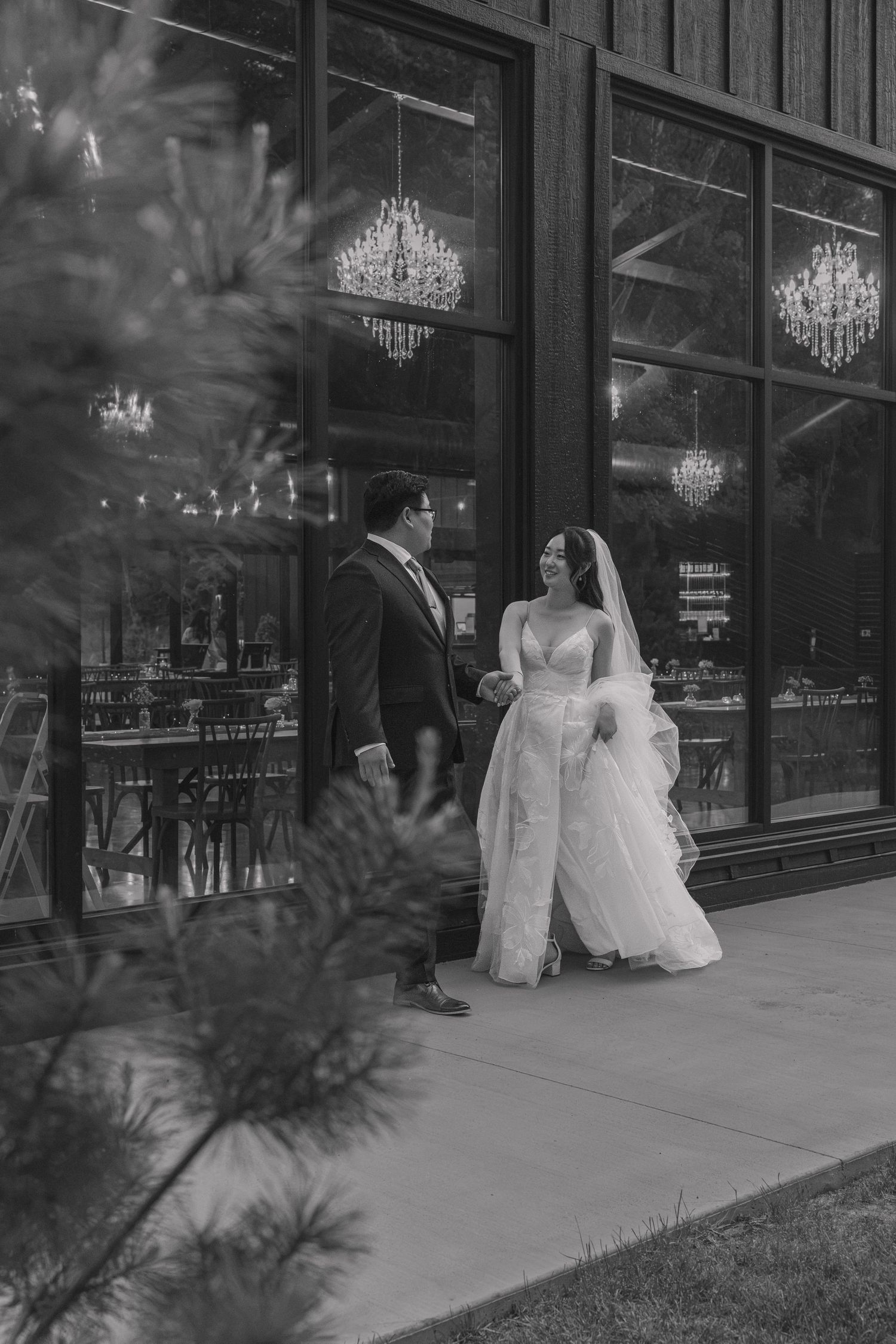 Black and white photo of bride and groom walking hand in hand outside elegant venue with chandeliers visible through large windows.