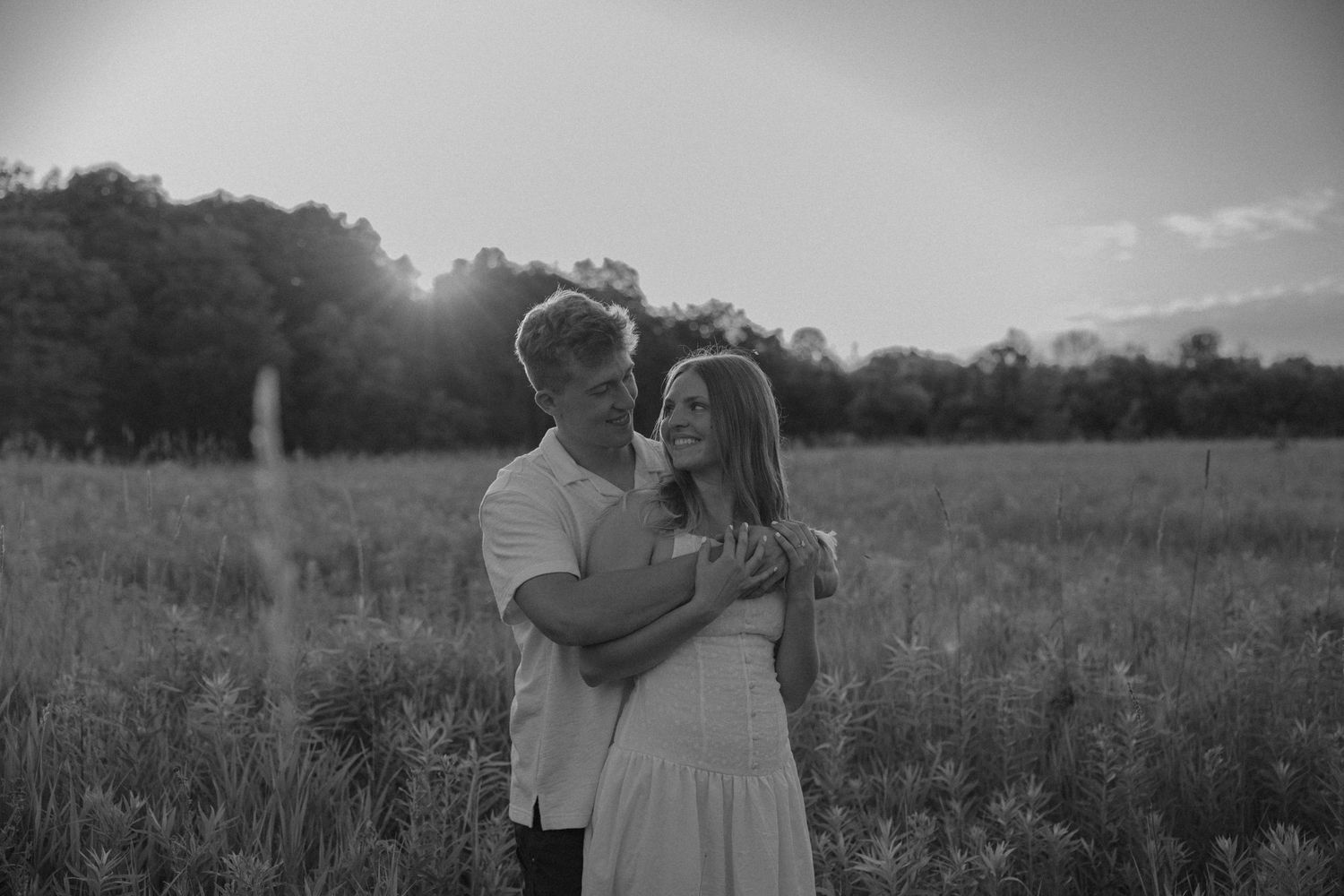 Black and white photo of a couple embracing tenderly in a field during golden hour.