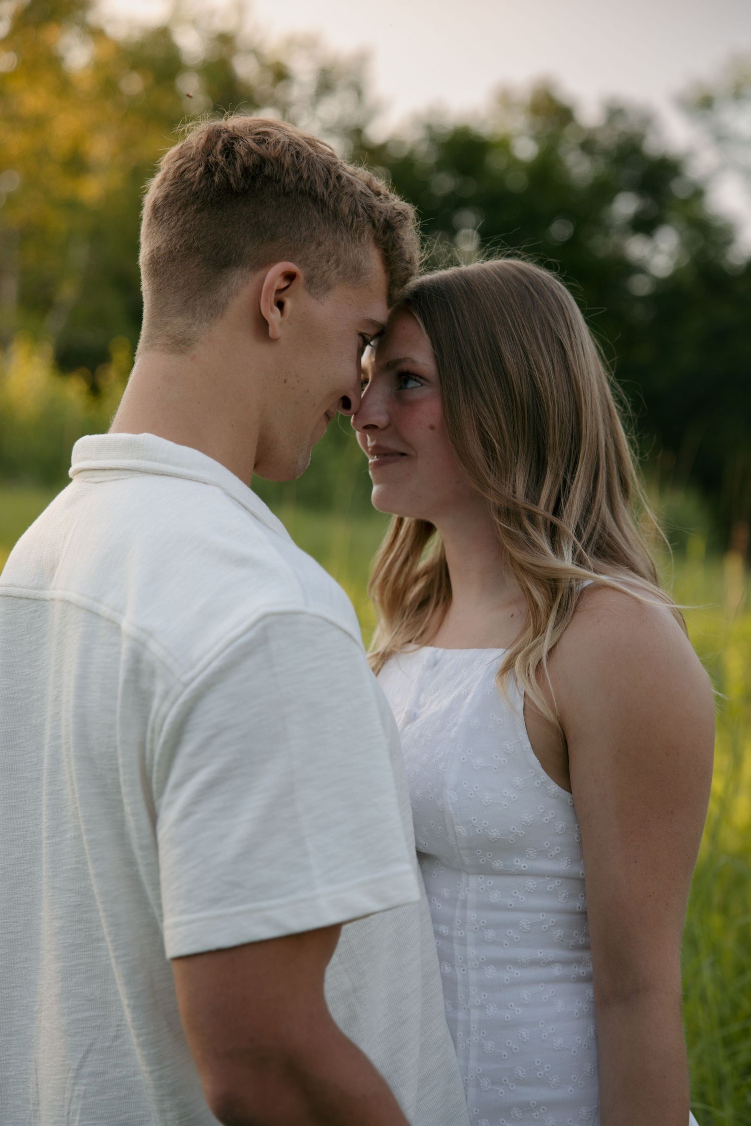 Young couple in white outfits sharing an intimate moment nose-to-nose in a sunlit green field.
