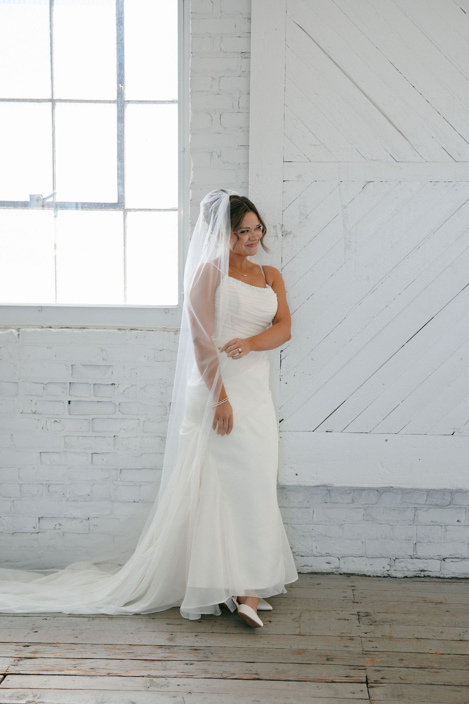 Elegant bride in a white satin gown with a long cathedral veil standing by a window in a white brick room.