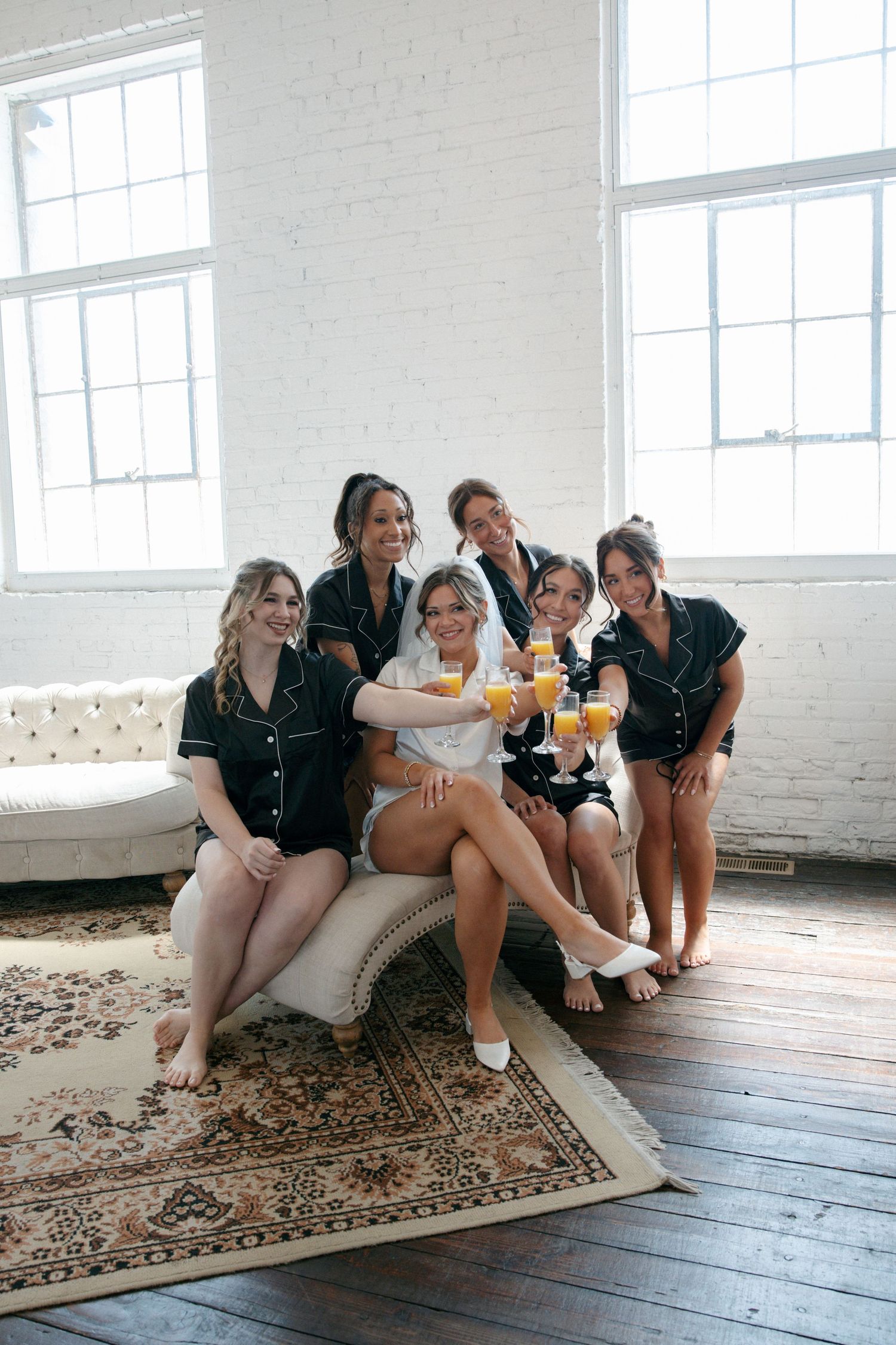 Bride and bridesmaids in black pajamas toasting with mimosas on tufted sofa before wedding.