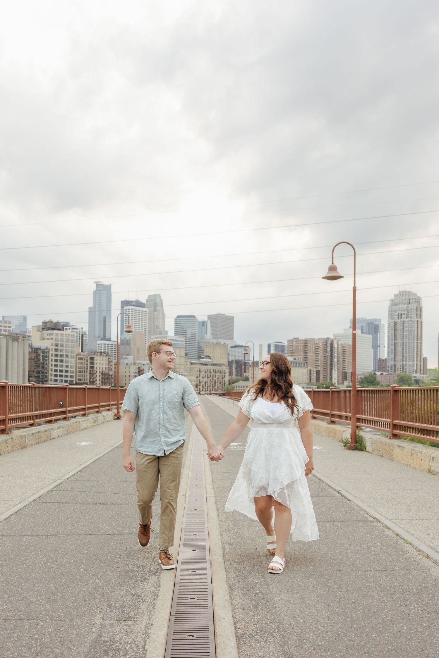 Couple holding hands walking on a bridge with Minneapolis city skyline in the background on a cloudy day.