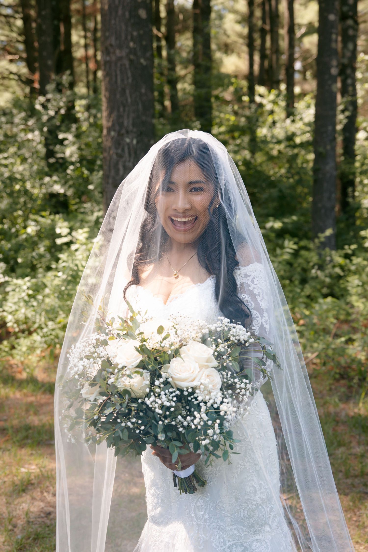 Smiling bride in white lace gown and veil holds bouquet of white roses and greenery in a sunlit forest.