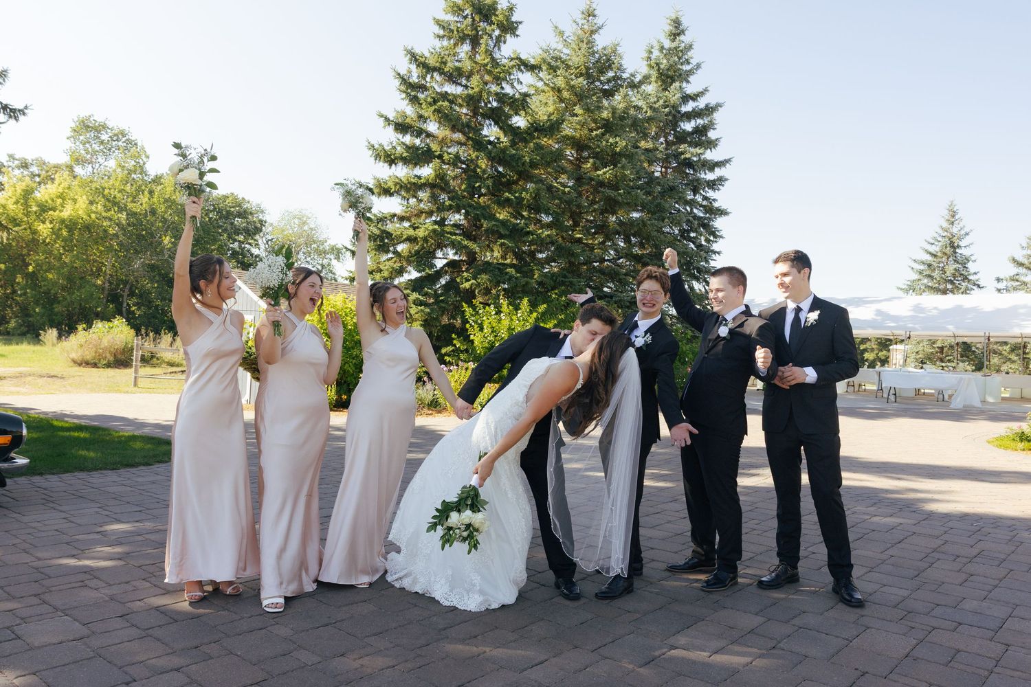 Wedding party celebrating as groom dips bride, bridesmaids in blush gowns and groomsmen in black suits outdoors.