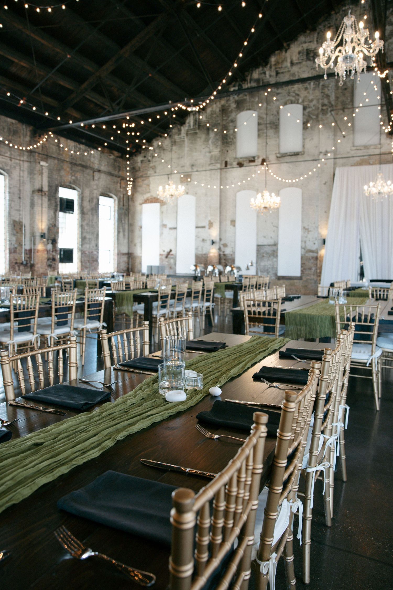 Industrial wedding reception hall with string lights, gold chiavari chairs, and green table runners.