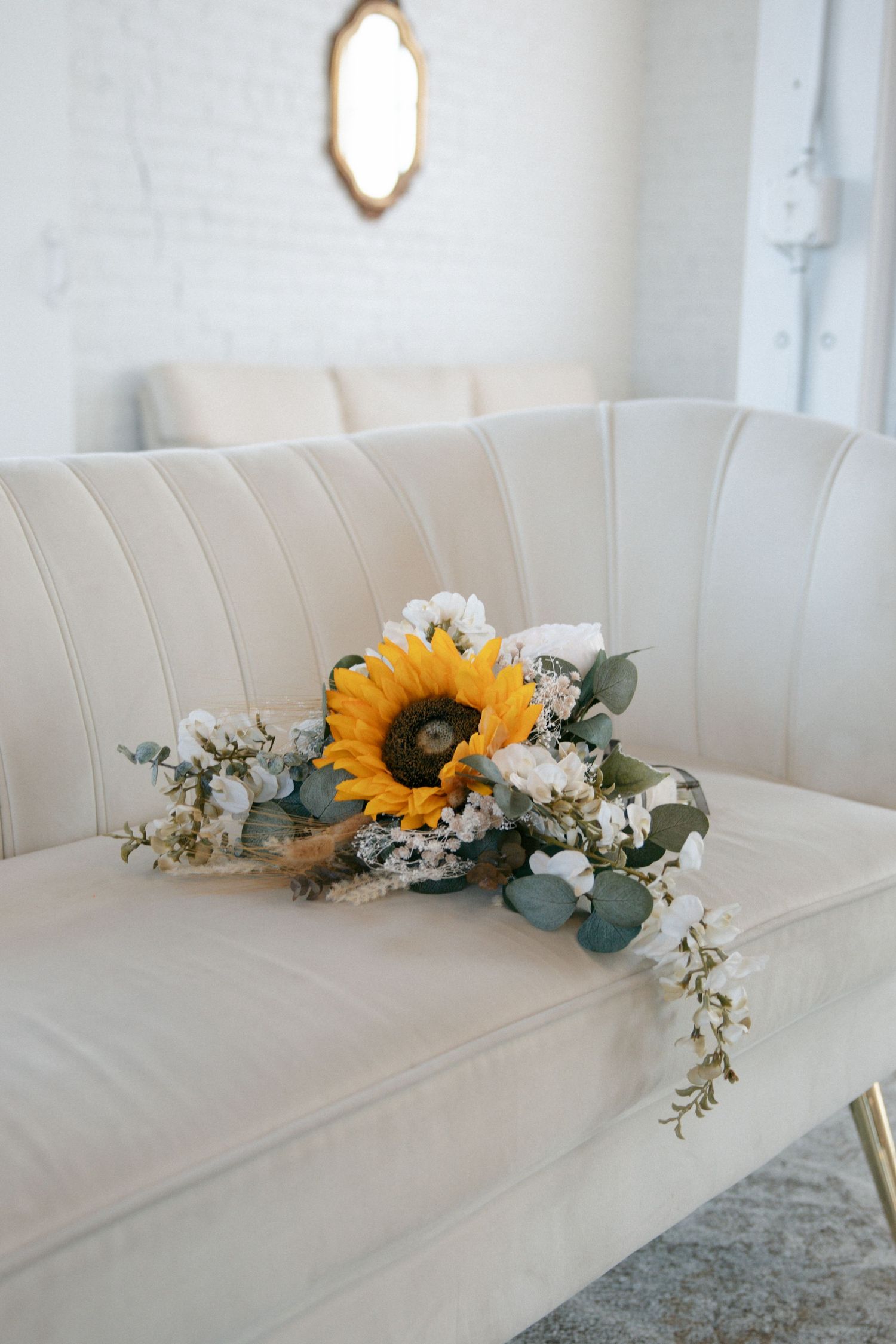 Sunflower bridal bouquet with white flowers and eucalyptus greenery resting on a cream velvet sofa.