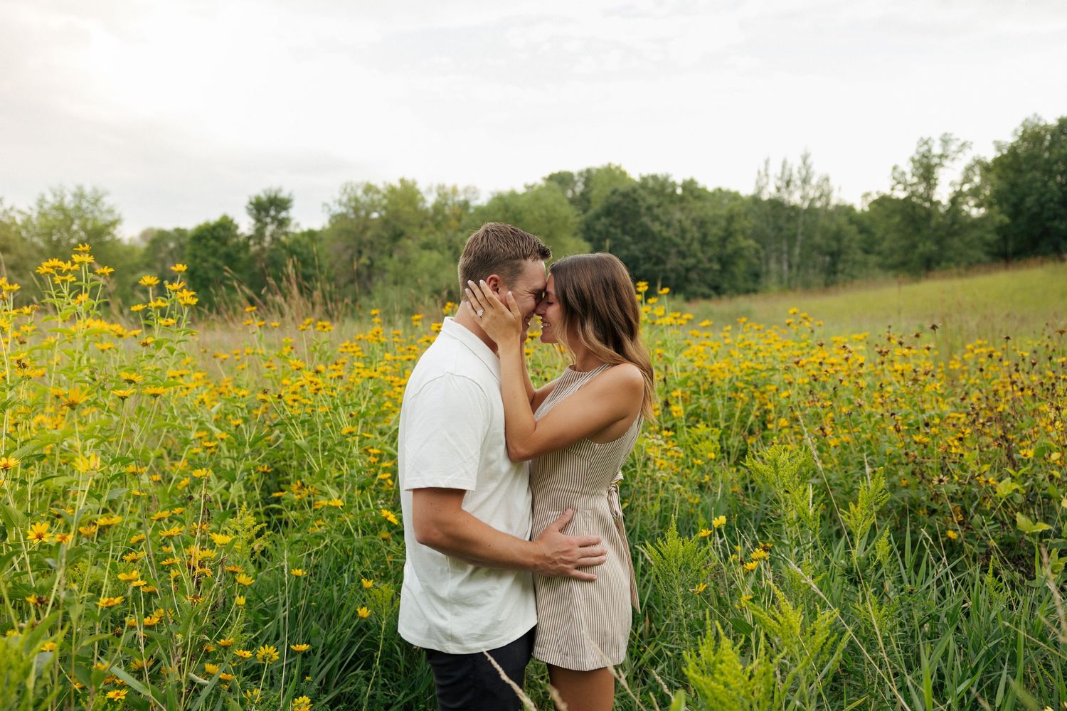 Couple sharing a romantic moment in a field of yellow wildflowers with lush green trees in background.