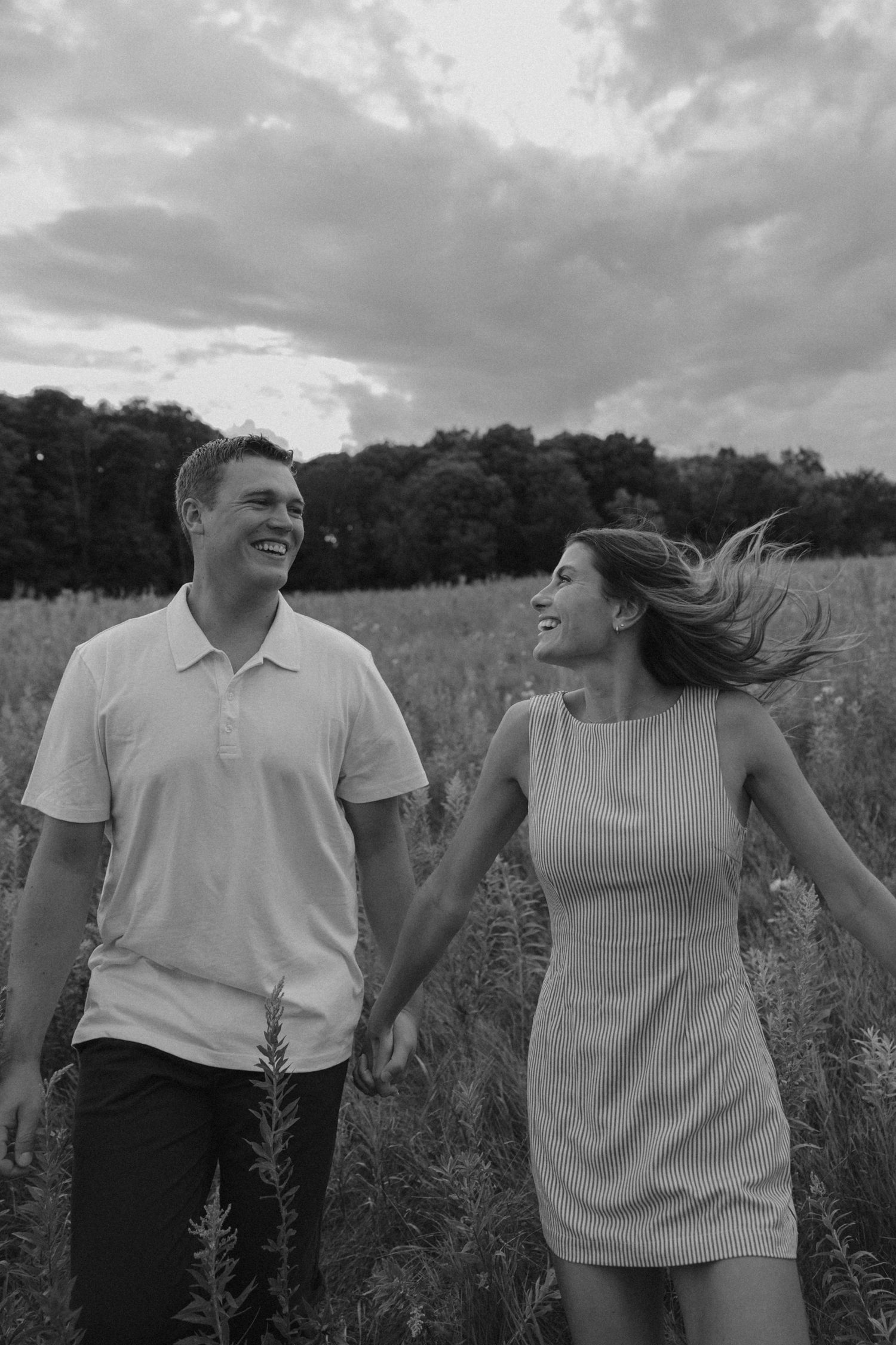 Black and white photo of a happy couple holding hands and laughing while walking through a field.