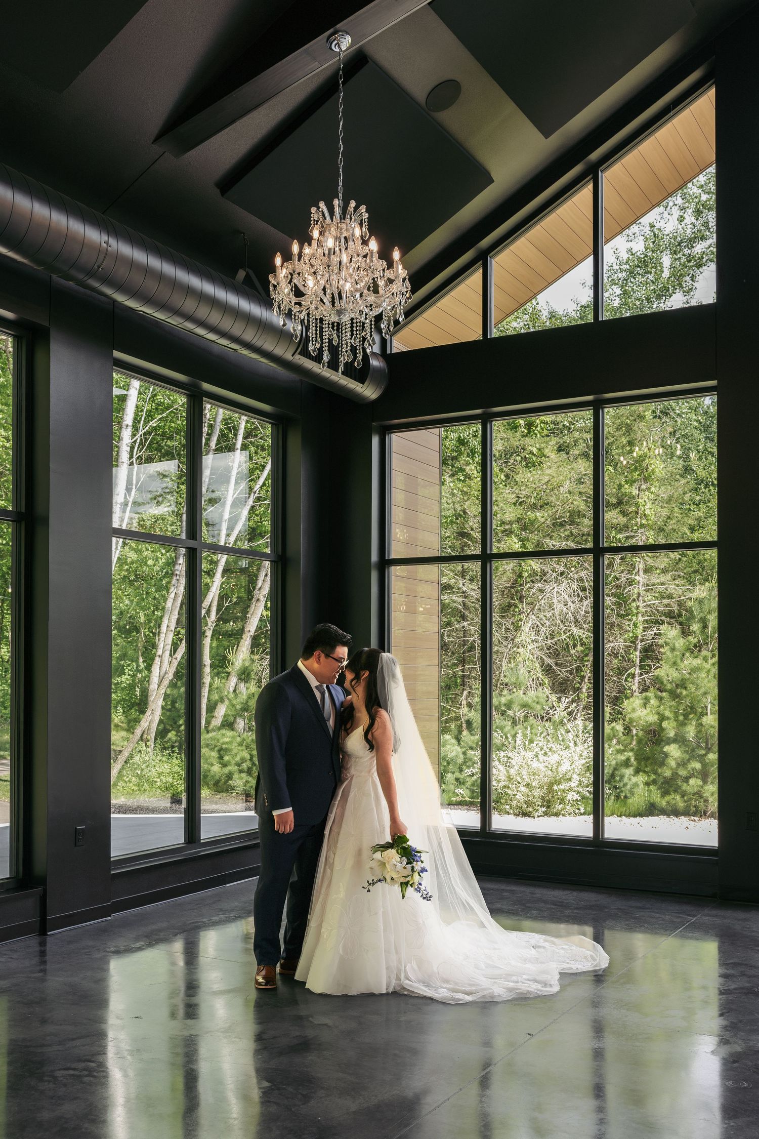 Bride and groom share a romantic moment under a crystal chandelier in a modern venue with floor-to-ceiling windows.