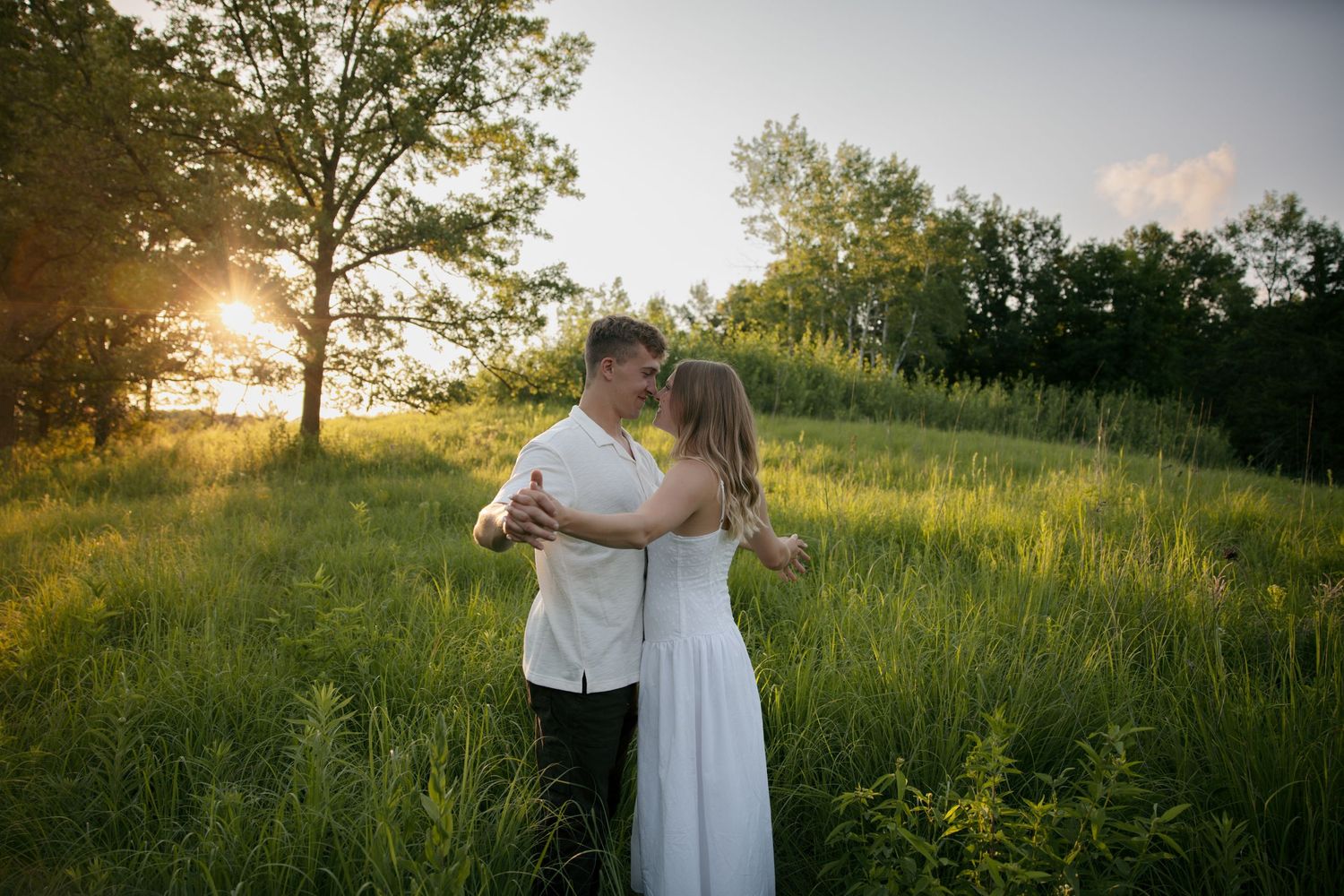 Couple embracing in a lush green meadow with golden sunset light streaming through trees.