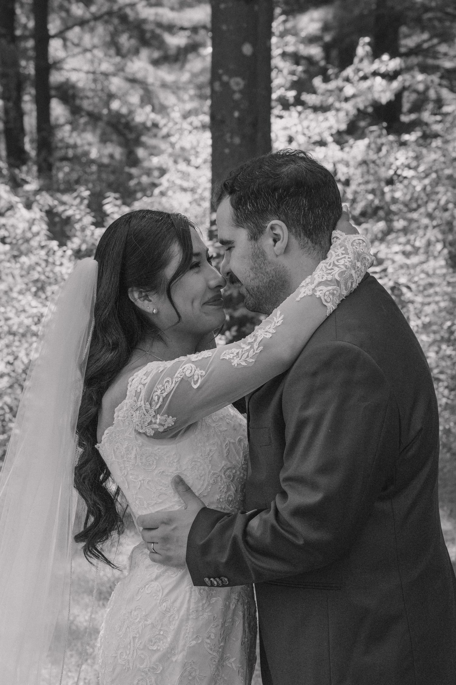 Black and white photo of a bride and groom sharing an intimate moment in a forest, wearing lace wedding dress and suit.
