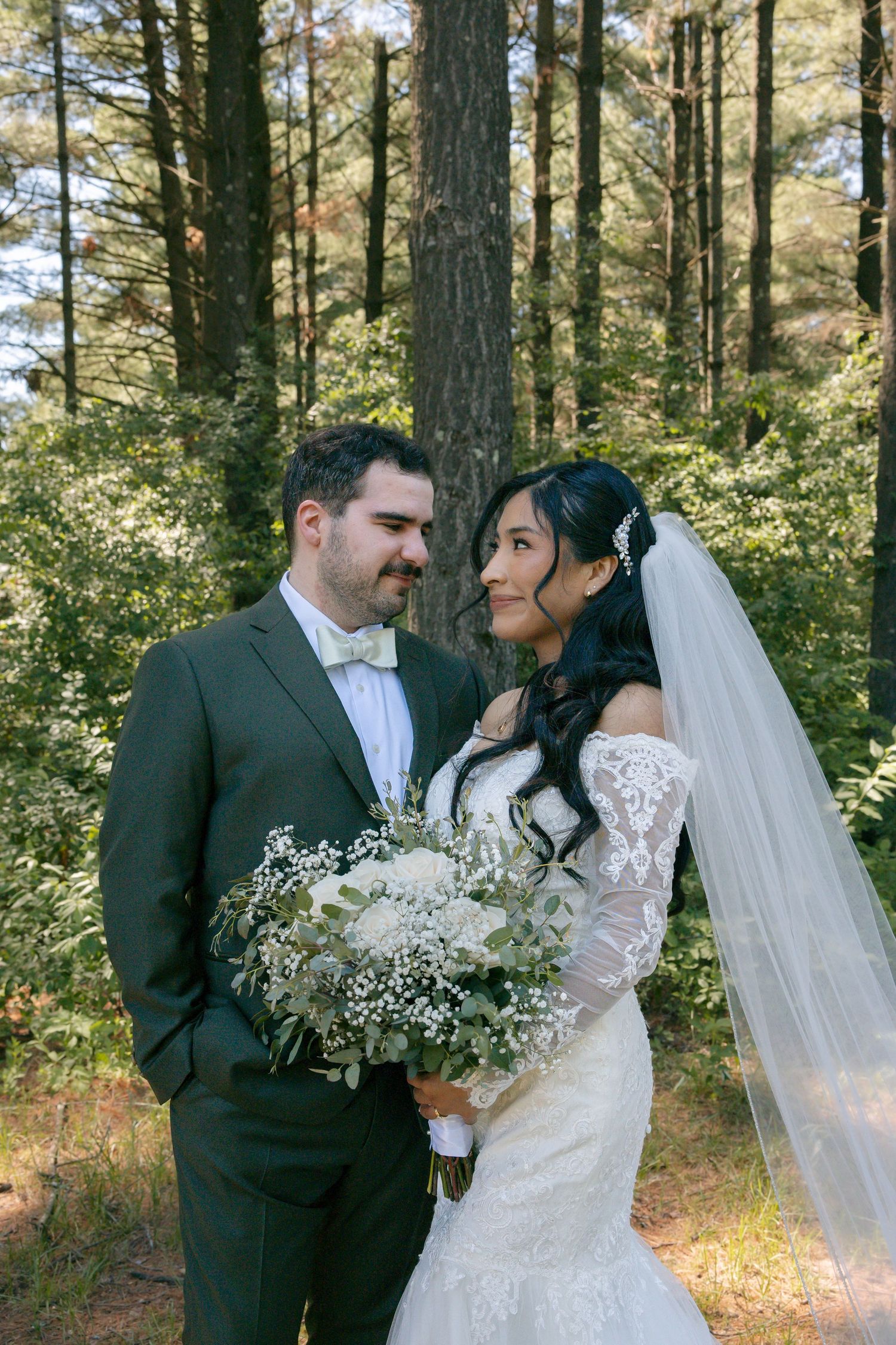 Smiling bride and groom share a tender look in a pine forest, she holds a white and greenery bouquet.