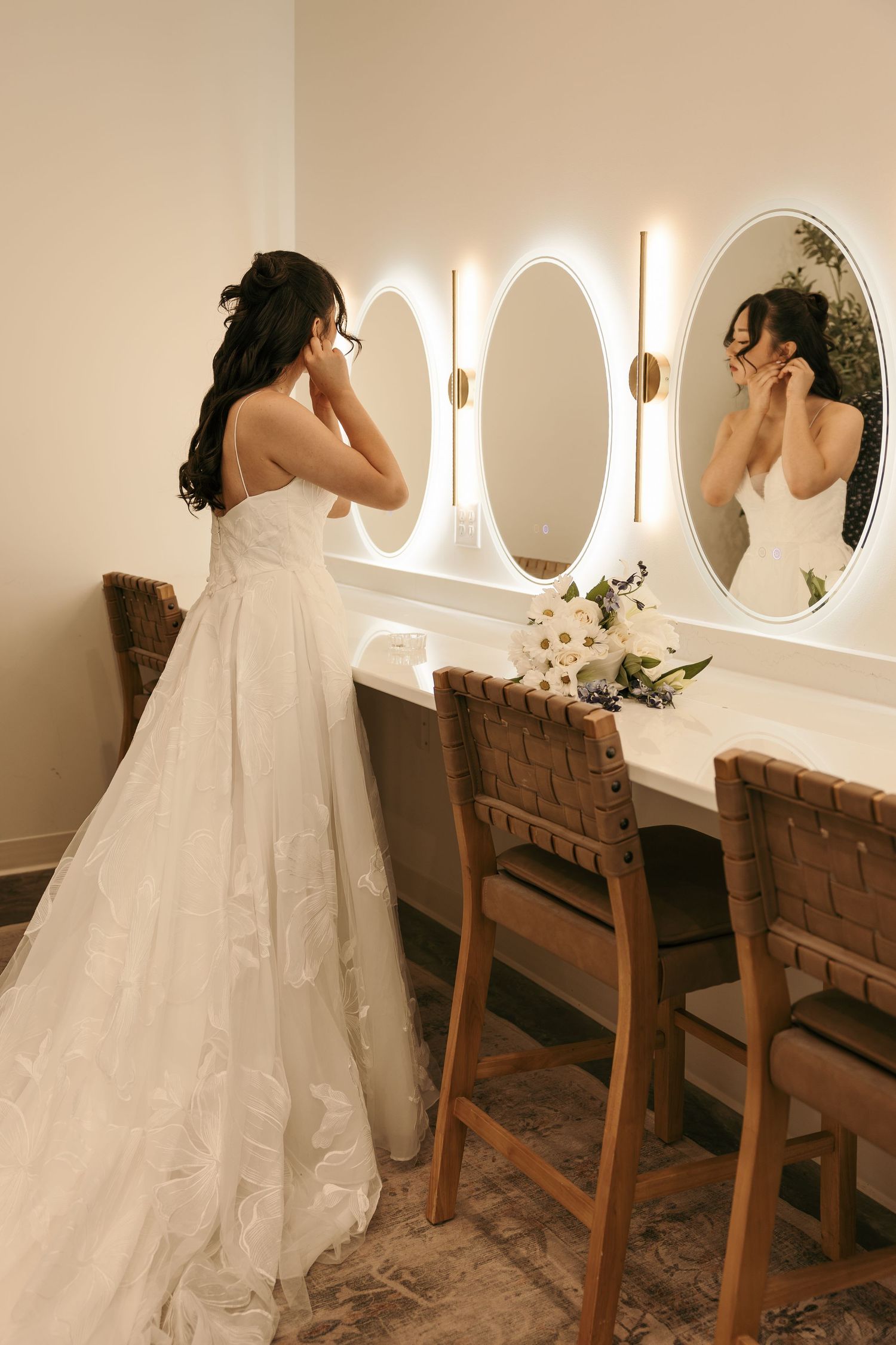 Bride in white gown adjusting earrings at vanity mirror with floral bouquet and warm lighting.