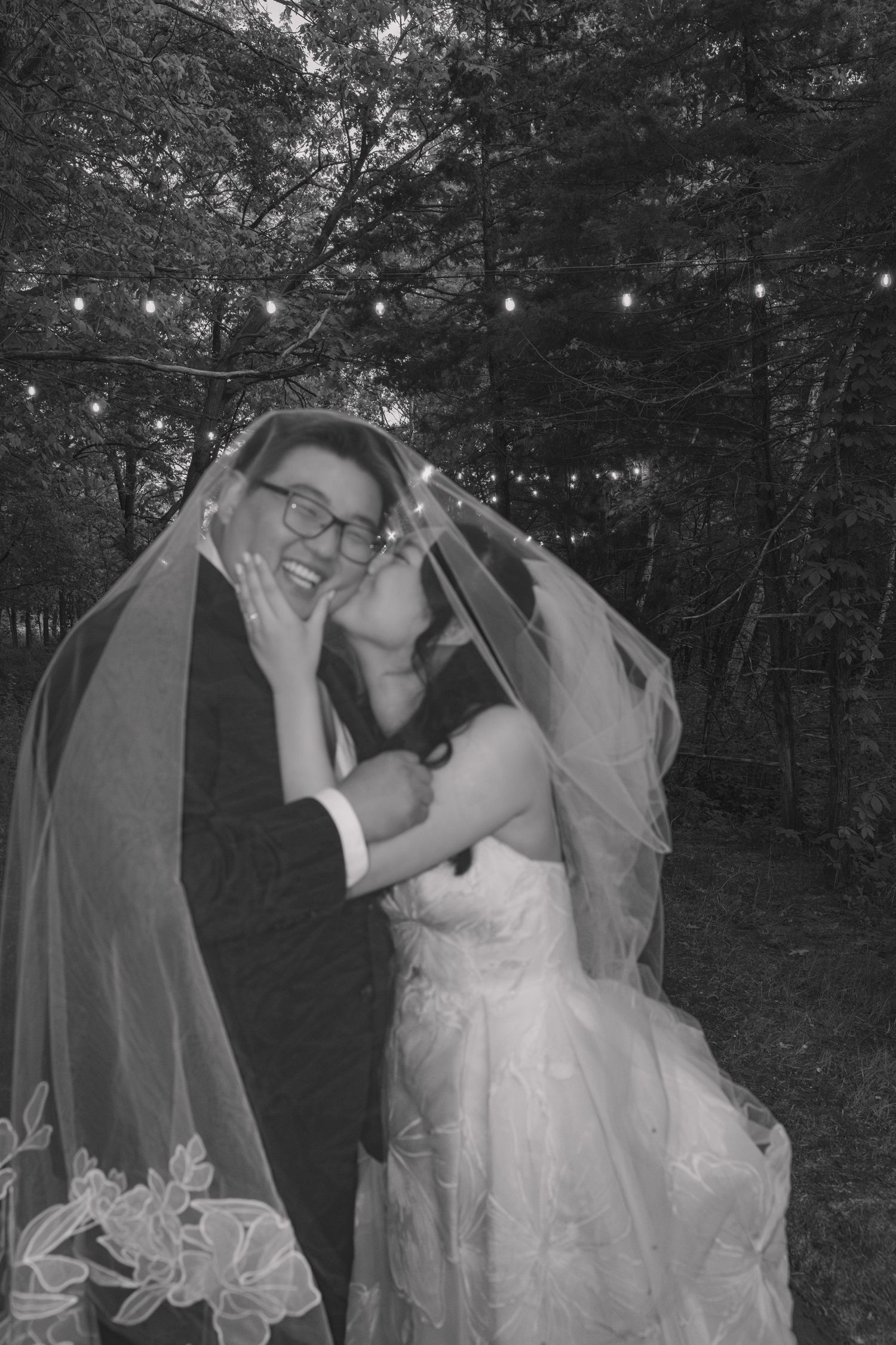 Black and white photo of a bride and groom sharing a romantic moment under her veil, surrounded by string lights in a forest.