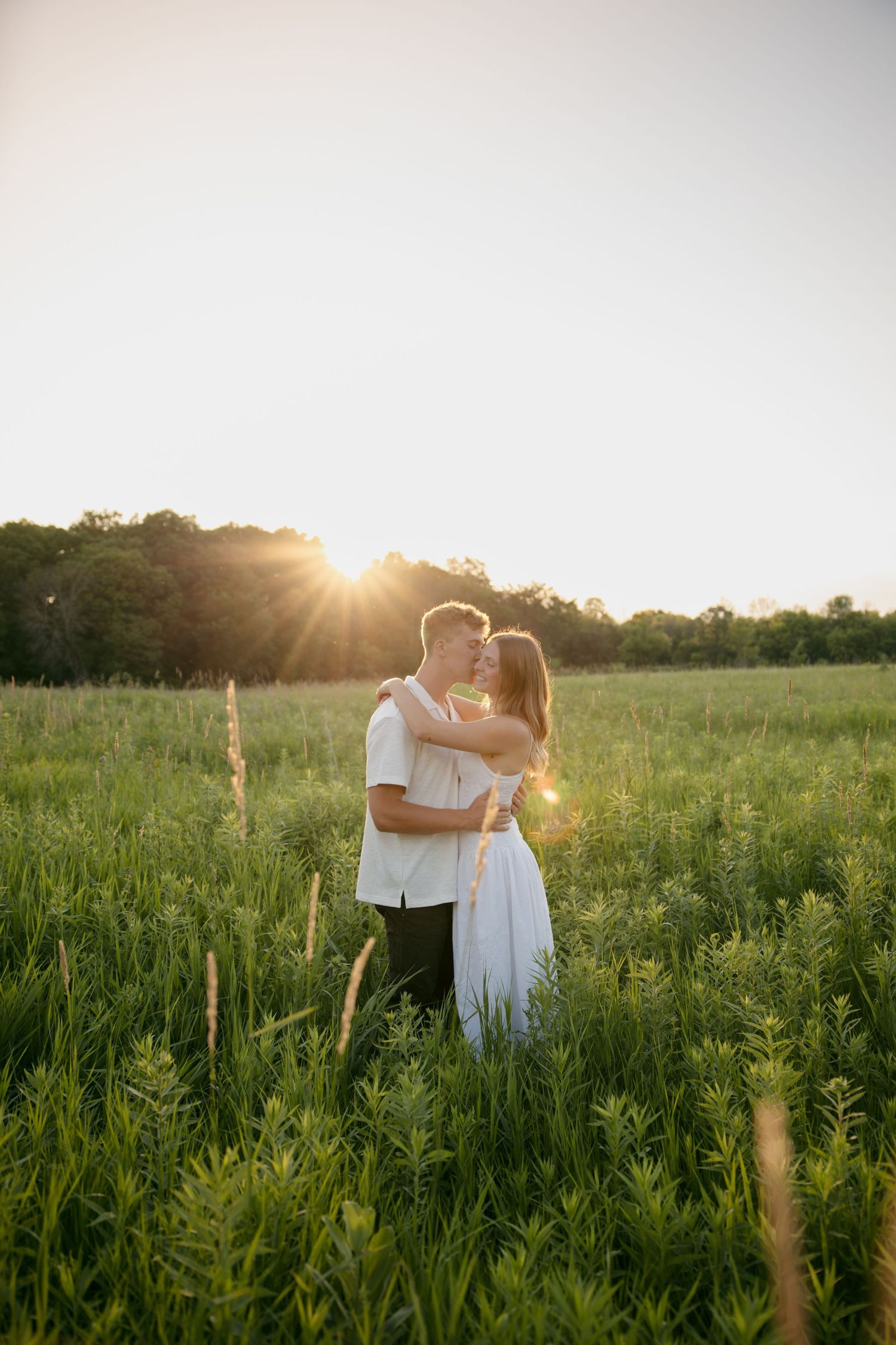 Romantic couple holding each other close in a sunlit green field at golden hour sunset.