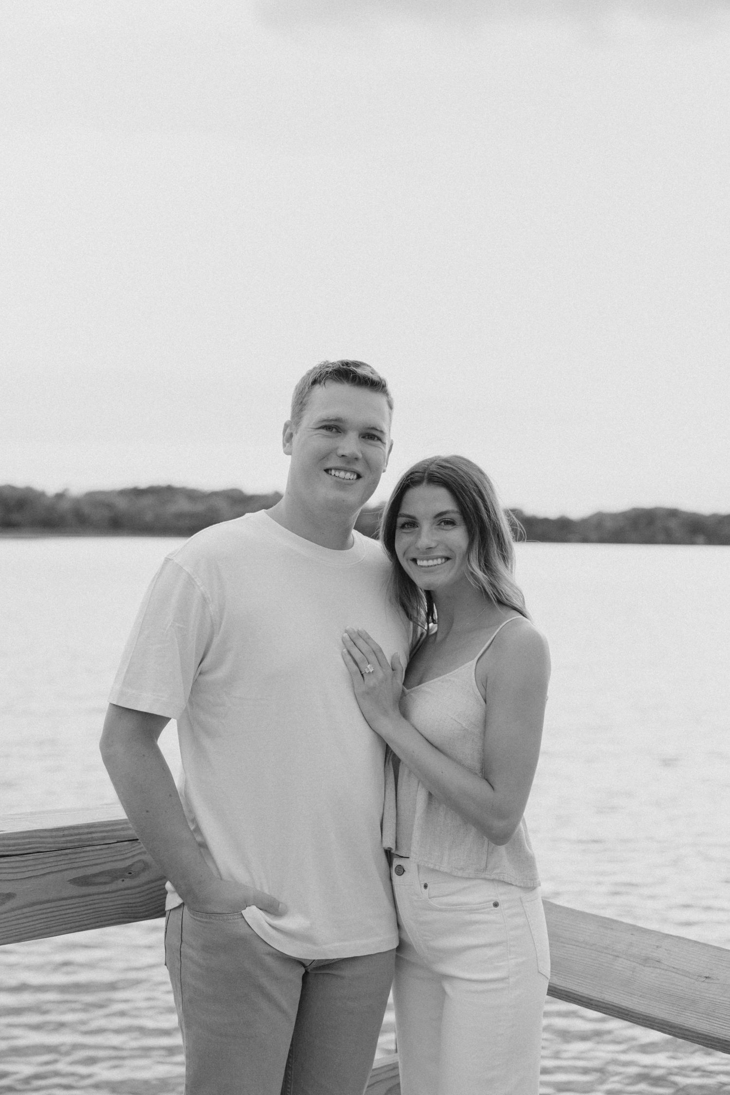 Black and white photo of smiling couple standing on a wooden dock by a calm lake.