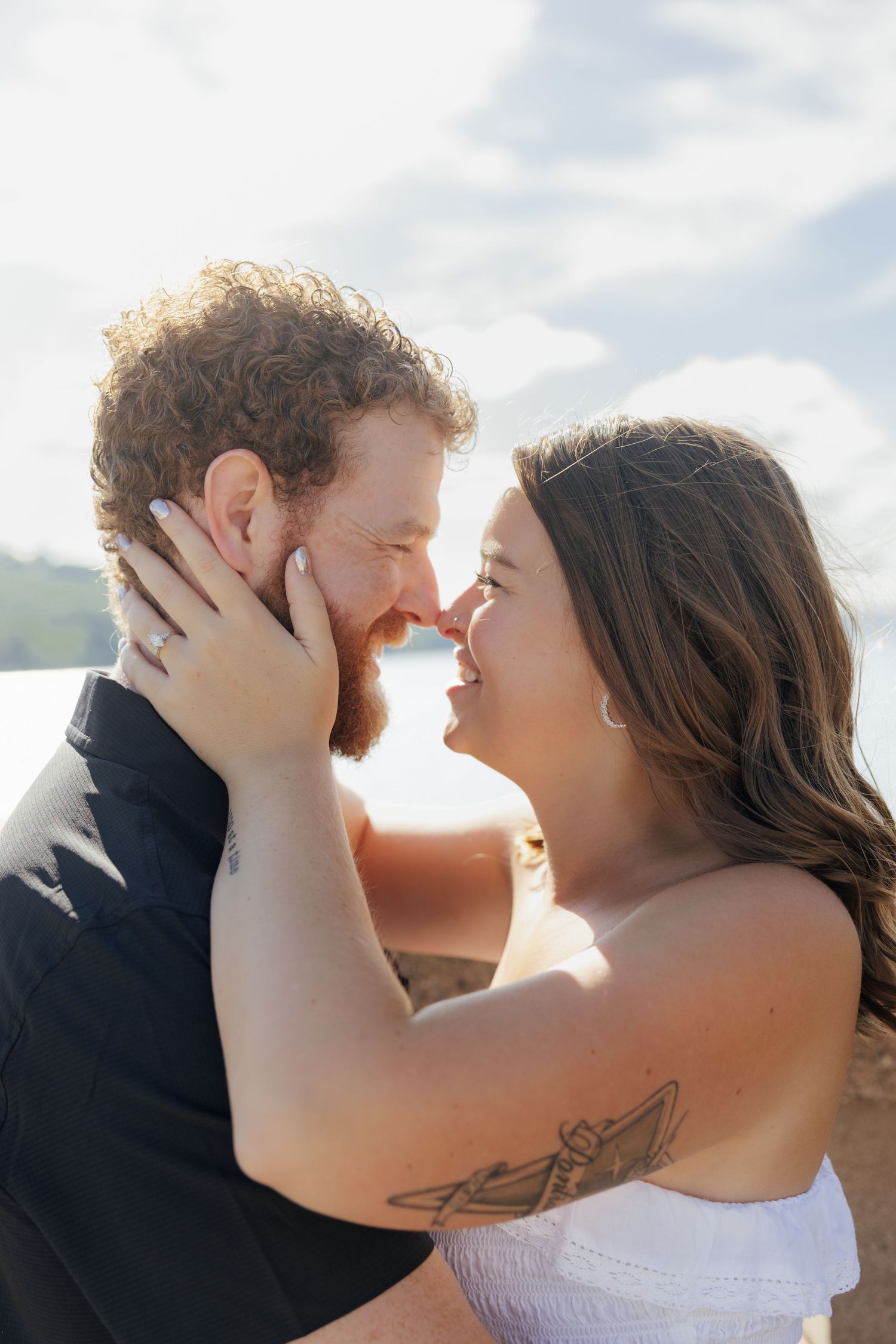 A couple shares a romantic almost-kiss outdoors, the woman in white holding the man's face, both smiling joyfully.