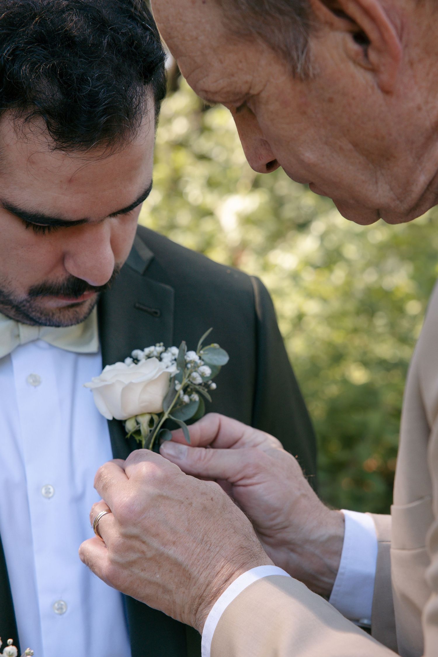 Older gentleman carefully pins a white rose boutonniere onto grooms dark suit lapel outdoors.