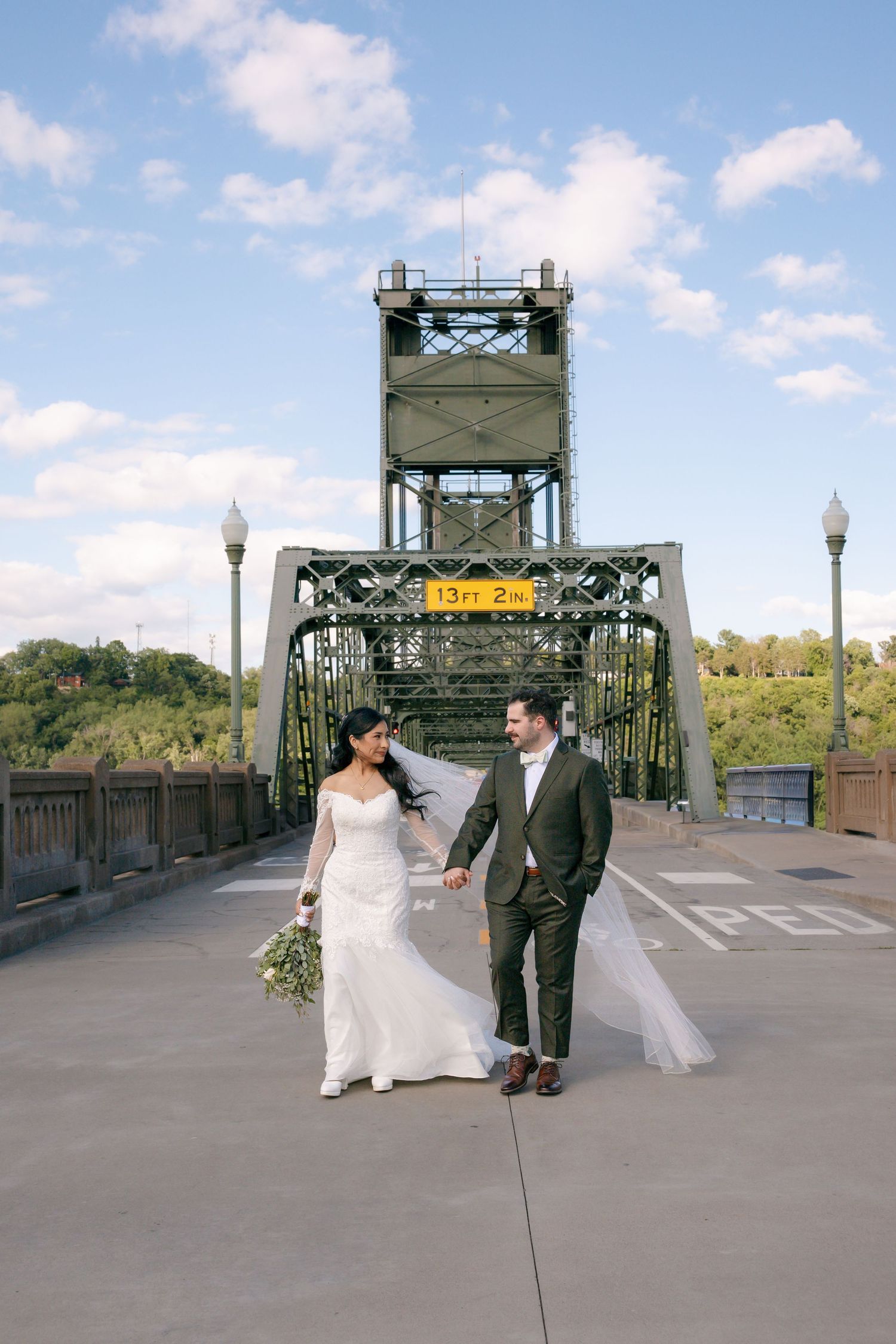 Bride and groom walk hand-in-hand on a bridge, her veil flowing, with a green steel lift bridge backdrop.