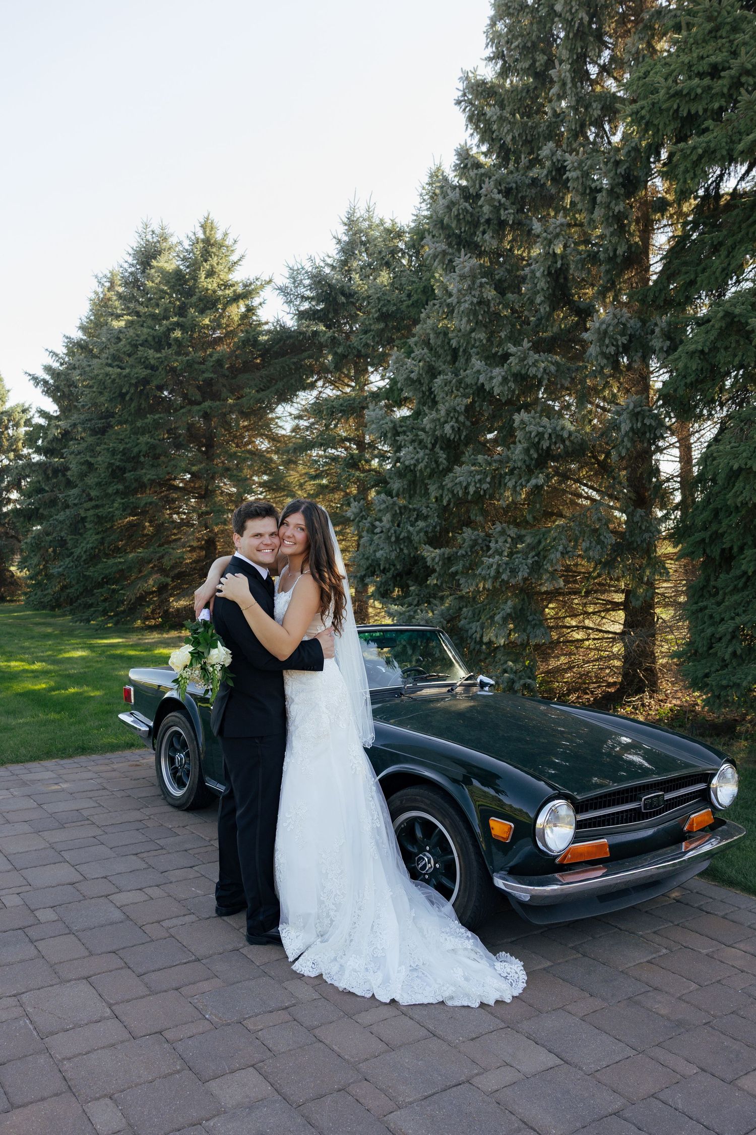Newlyweds embracing in front of a classic dark green convertible car on a brick driveway with evergreen trees.